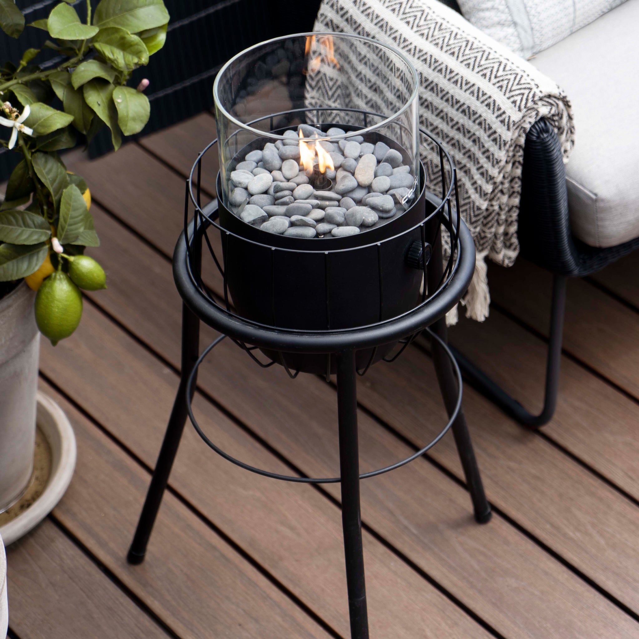 A Cosi - Cosiscoop Basket High Black Lantern with stones on a wooden deck near a plant and woven blanket offers a modern chic outdoor light source.