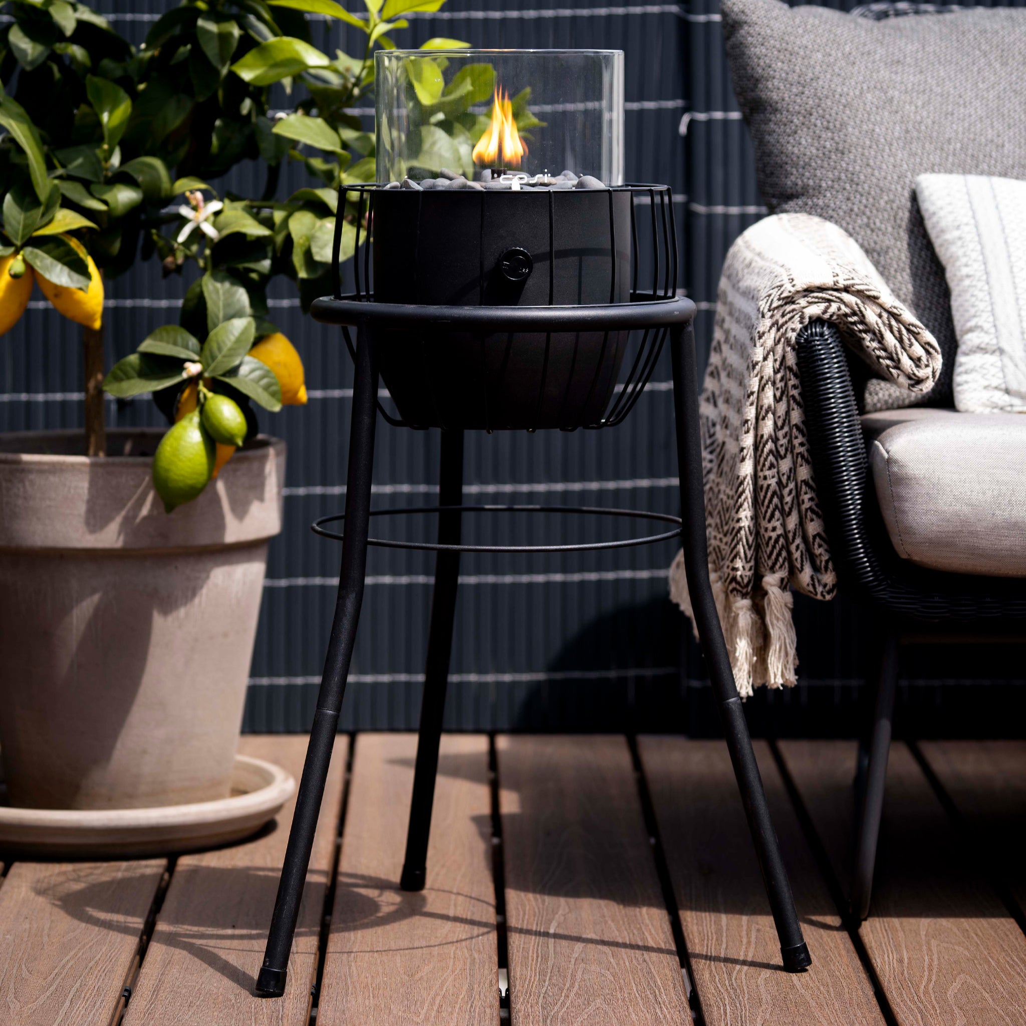 A small fire pit flickers next to a potted lemon tree and a cushioned chair on the wooden deck, enhanced by the Cosi - Cosiscoop Basket High Black Lantern, adding modern chic as an outdoor light source.