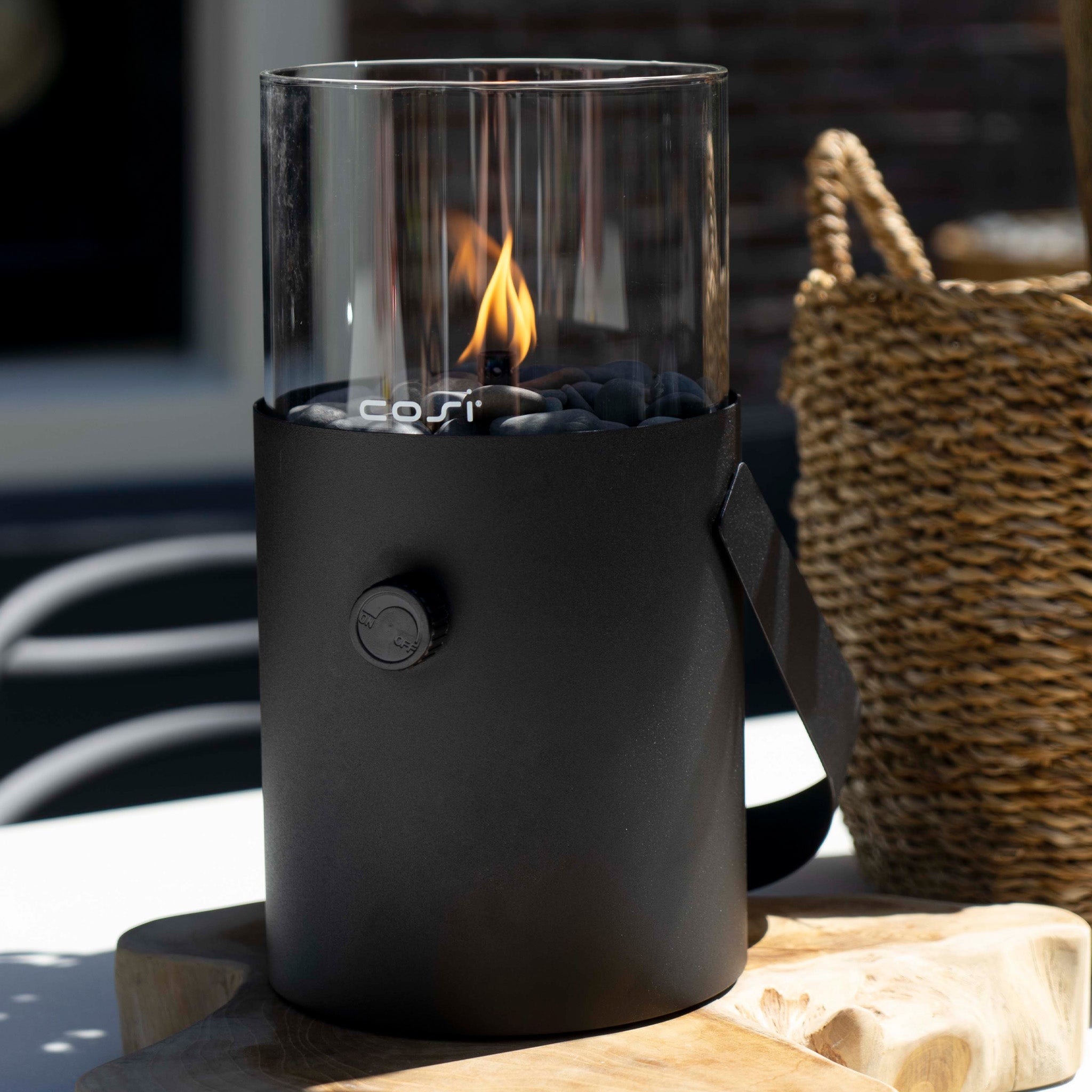 The Cosi - Cosiscoop Fire Lantern in Black is a modern portable lantern that flickers with a visible flame, casting a warm glow on wood beside a woven basket—ideal for outdoor lighting.