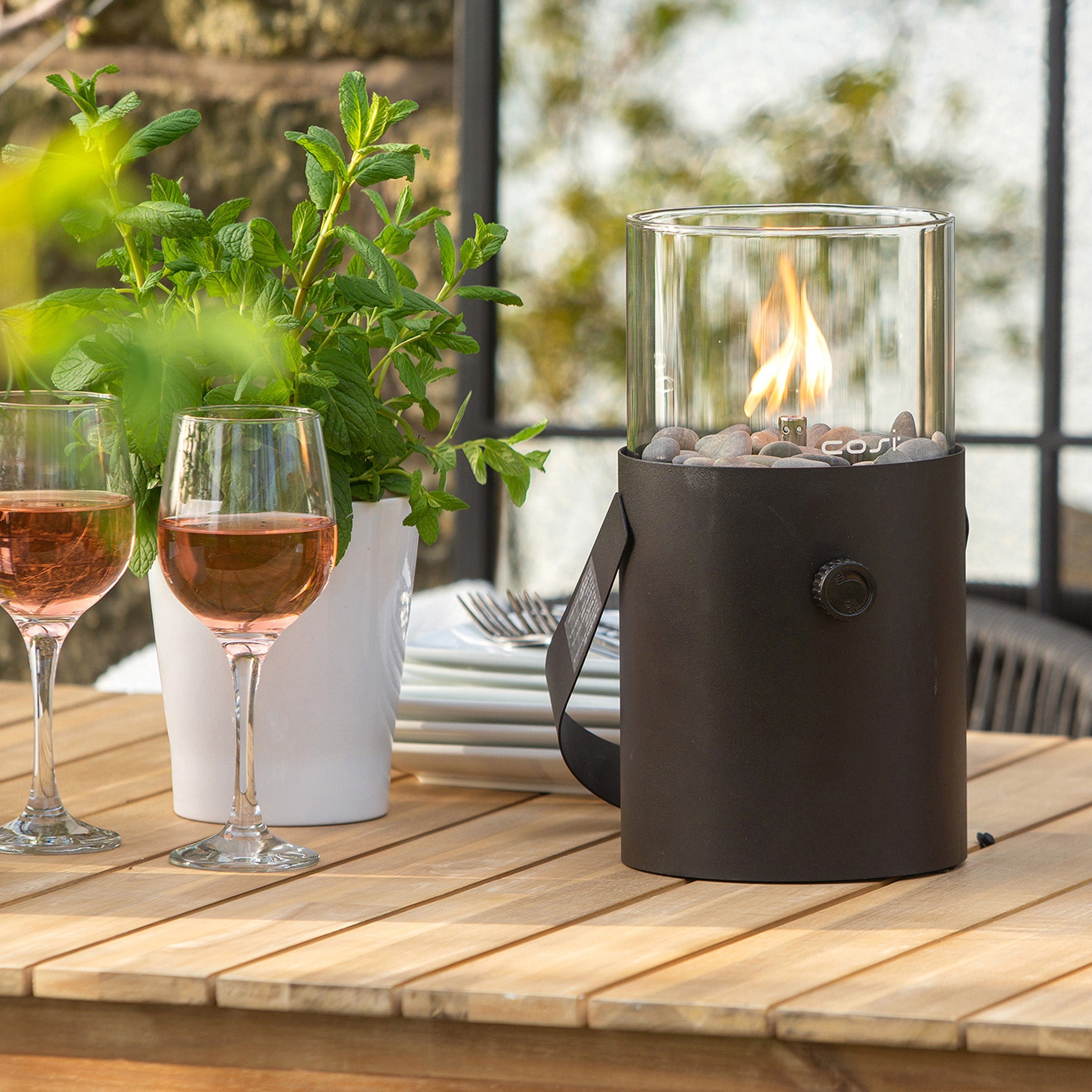 Table adorned with a Cosi - Cosiscoop Fire Lantern in Black, two glasses of rosé wine, and a potted plant, ideal for cozy outdoor evenings.