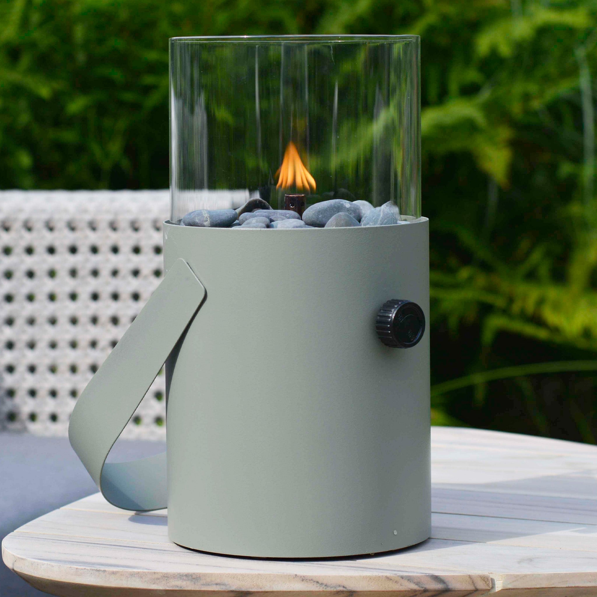 The Cosi - Cosiscoop Fire Lantern in Green, with its small tabletop design and glass shield, complements a garden lantern perfectly on a light wood outdoor table.