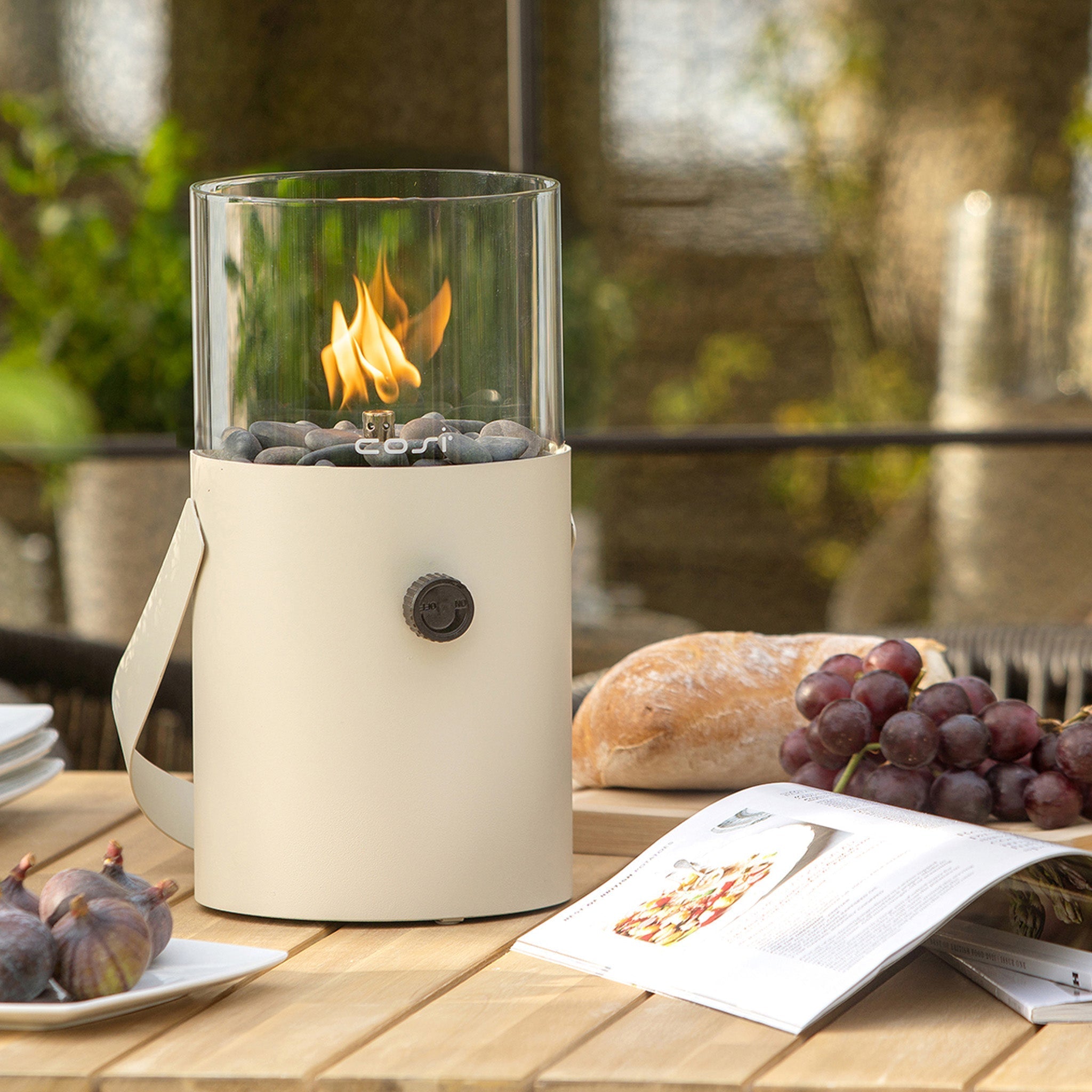 A Cosi - Cosiscoop Fire Lantern in Ivory casts a warm glow over a wooden table with grapes, a baguette, figs, and an open menu, creating a cozy outdoor setting.