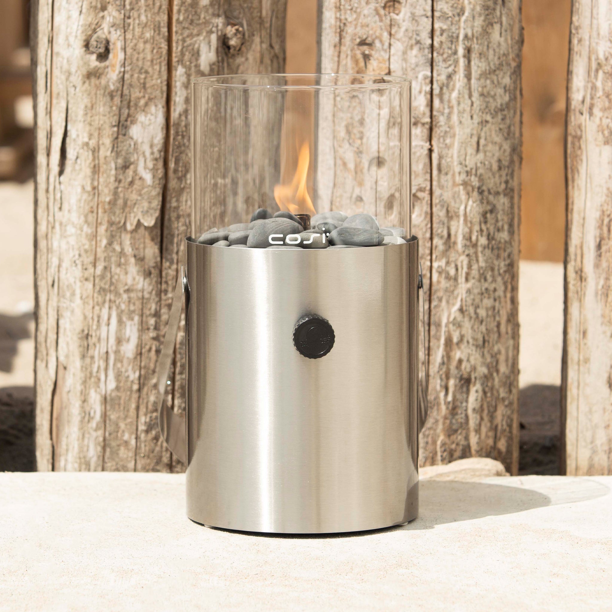 The Cosi - Cosiscoop Fire Lantern in Stainless Steel, with black stones and a small flame, captures the charm of an outdoor fire lantern on wooden planks.