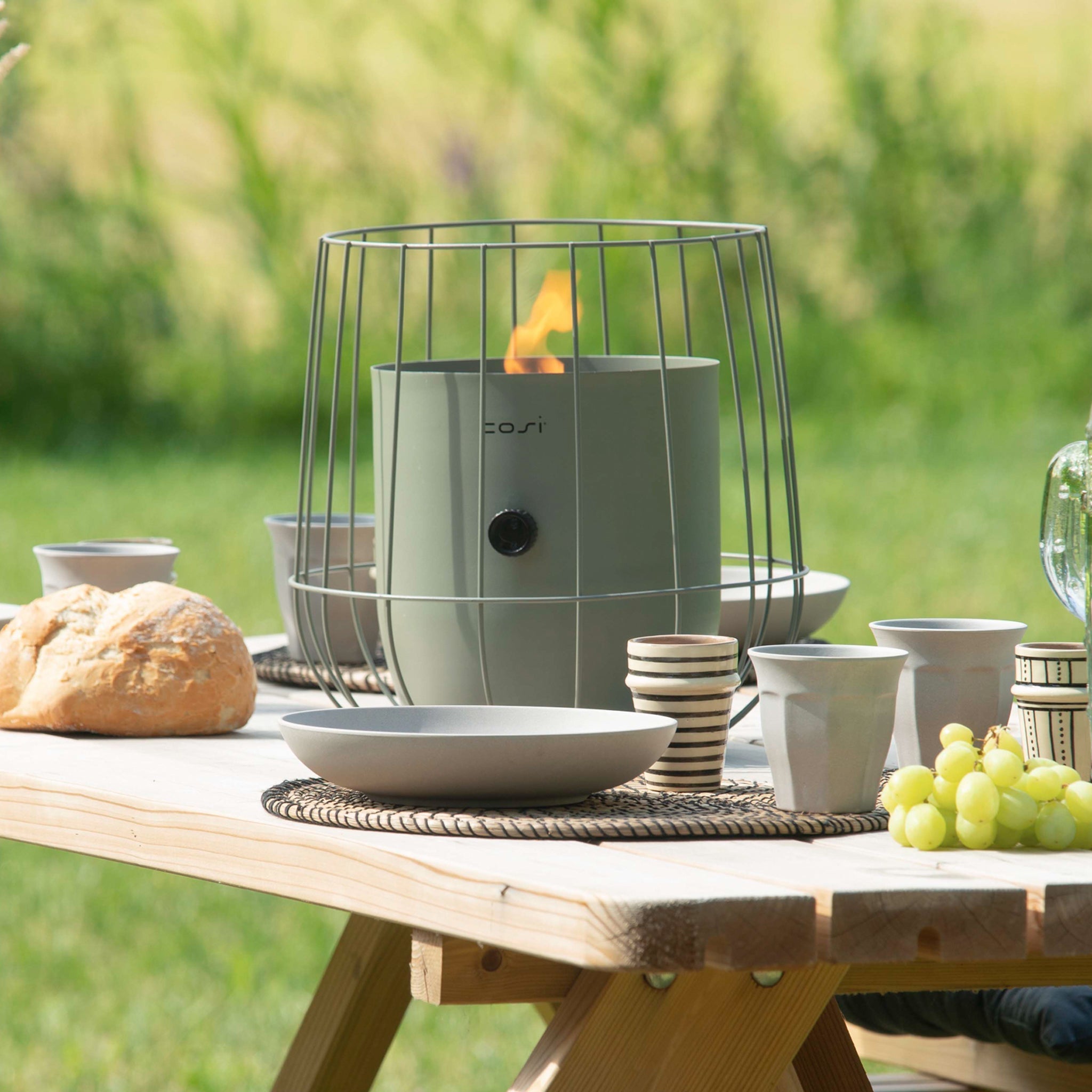 A picnic table with dishes, bread, grapes, and a small heater in a wire frame is set outdoors in a green field, illuminated by the warm glow of the Cosi - Cosiscoop Basket Lantern in Olive.
