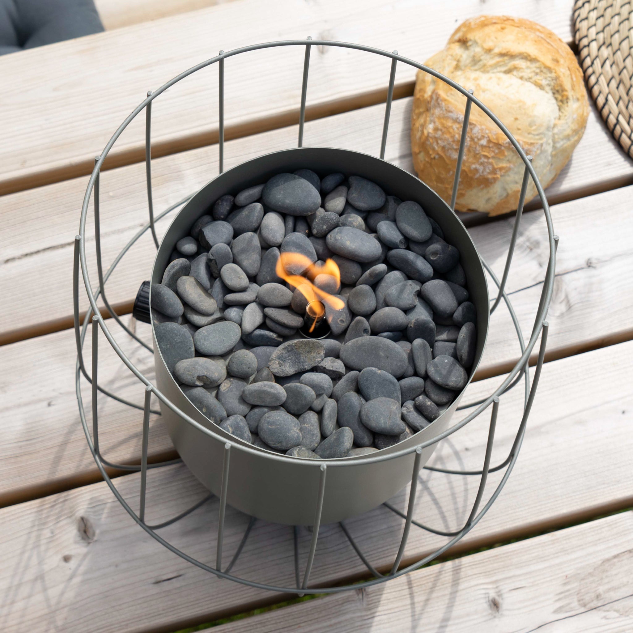 The Cosi - Cosiscoop Basket Lantern in Olive, with its stones and flame, sits on a wooden table beside bread, creating a cozy outdoor light source.