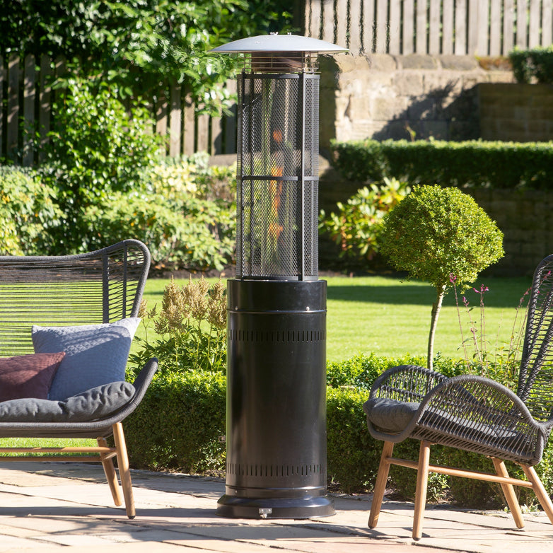 The Pacific Lifestyle Cylinder Patio Heater in black stands tall between two wicker chairs on a sunny patio, its real flames warmly dancing, powered by a propane gas canister, with a thriving garden in the background.