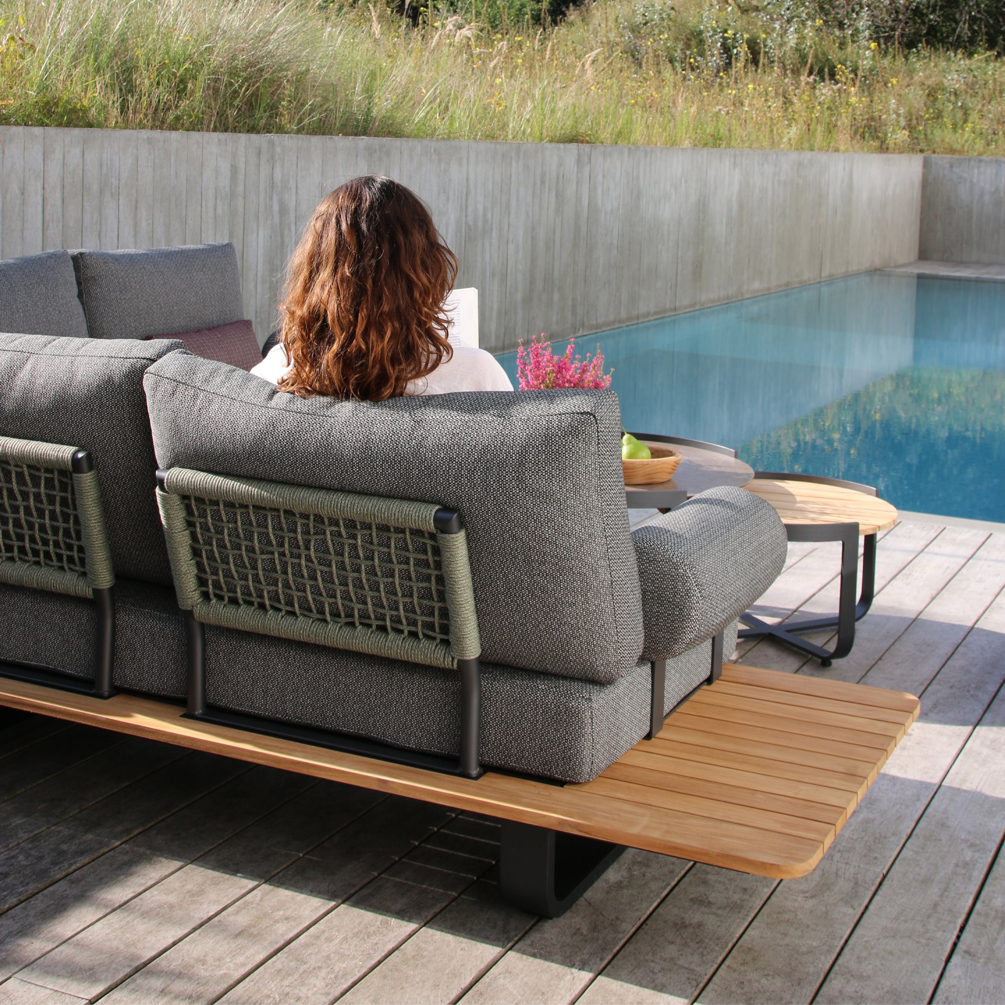 A person with wavy brown hair is sitting on a Bay Large Corner Group Set with 4 Waist Pillows and Teak Table in Olive Green beside a pool, reading a book. Green grass and a concrete wall are in the background.