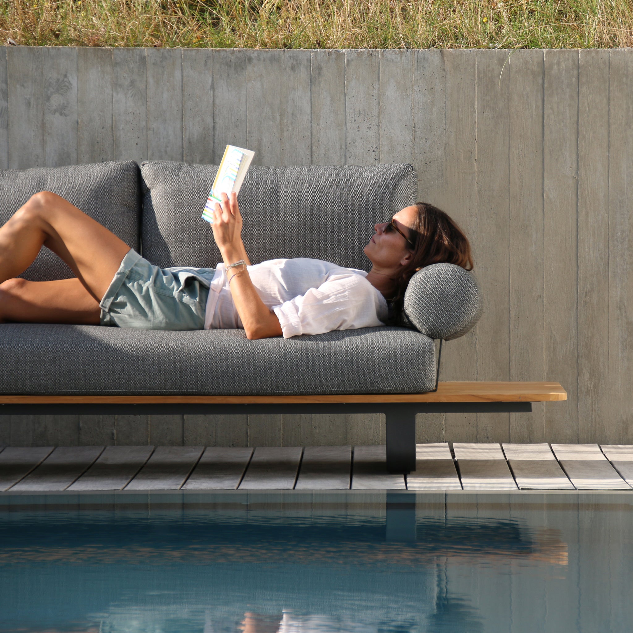 A woman enjoys her time reading a book on a Bay Corner Group Set with 4 Waist Pillows in olive green by the pool, the intricate acrylic rope design adding a touch of elegance. She lounges comfortably in the sun, wearing sunglasses, while a teak table beside her holds a chilled drink within easy reach.