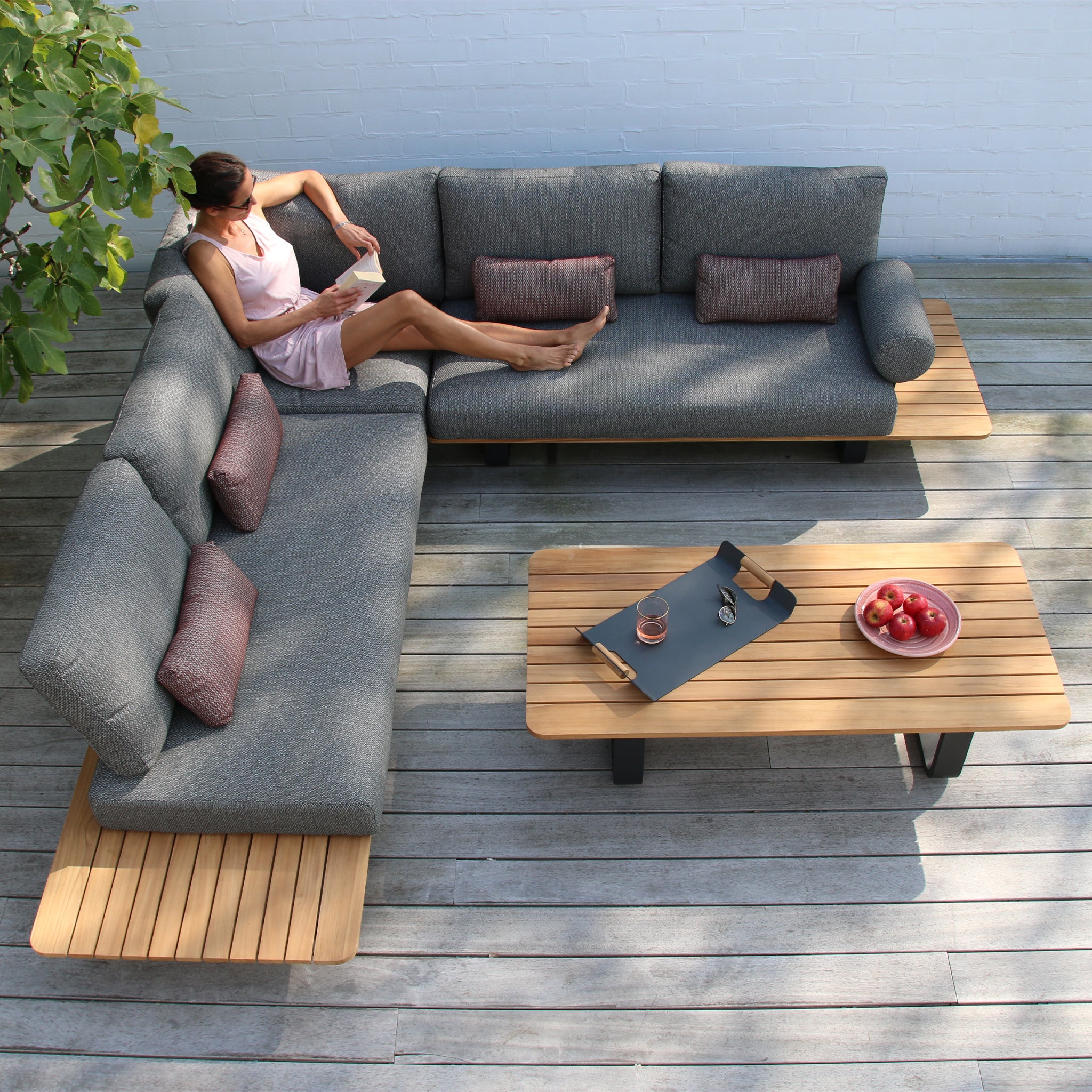 Person reclining on a modern L-shaped grey Bay Corner Group Set with 4 Waist Pillows in Olive Green on a wooden patio, with a teak table holding a tray and a bowl of apples.