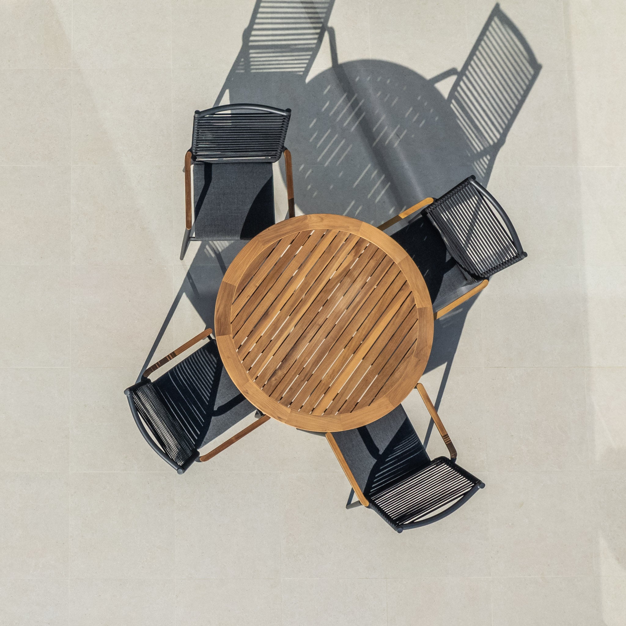 The Portland 4 Seat Round Dining Set with a Teak Table in Charcoal, featuring four black flat rope weaved armchairs, casts elegant shadows on a light-colored floor when viewed from above.