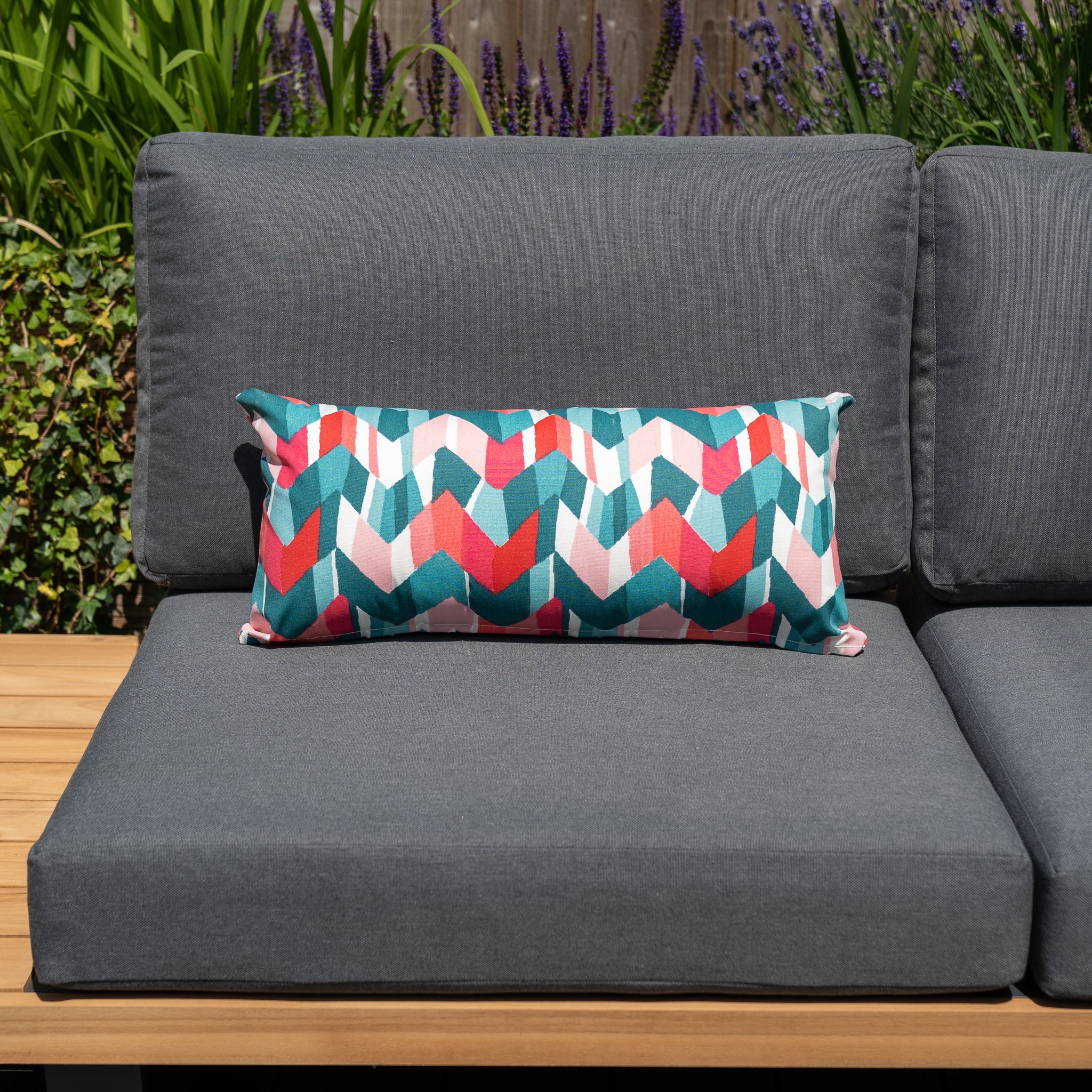 A gray outdoor sofa with a colorful chevron-patterned pillow on a wooden deck, featuring plants in the background. The arrangement is complemented by the vibrant Acrisol Flash Multi Bolster Scatter Cushion (60cm x 30cm), made from solution-dyed acrylic for long-lasting color.