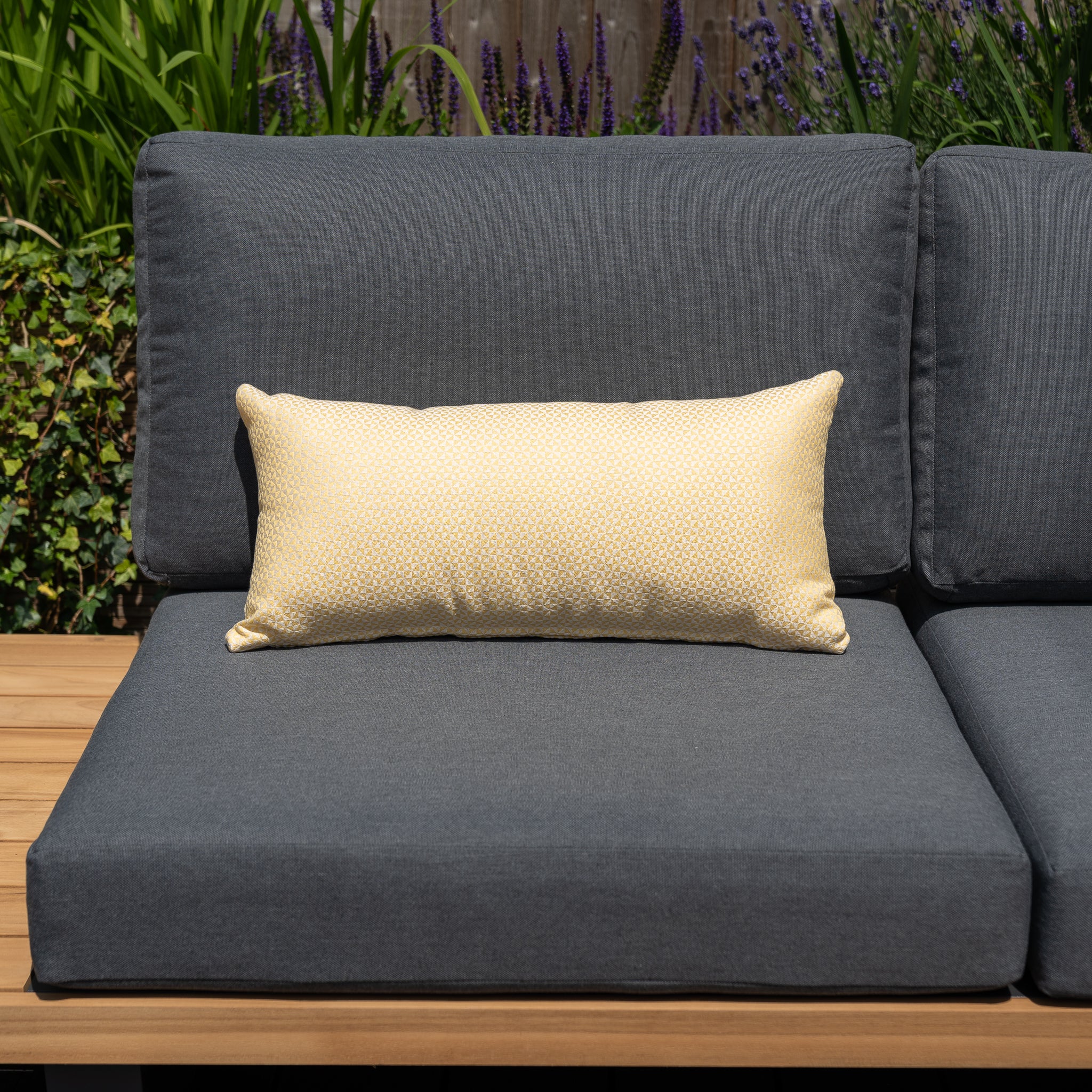 Outdoor gray cushioned seat featuring Harbour Lifestyle cushions with an Acrisol Helix Trigo Bolster Scatter Cushion (60cm x 30cm) in yellow, surrounded by greenery and purple flowers in the background.