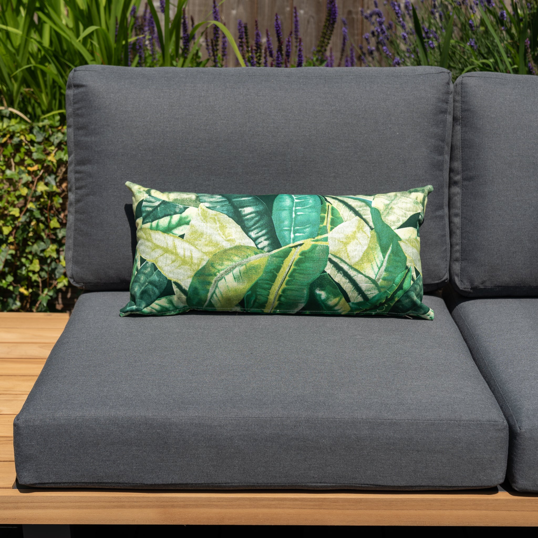 Gray outdoor couch with Acrisol Amazonia Verde Bolster Scatter Cushions, set against a backdrop of lush green plants and purple flowers.