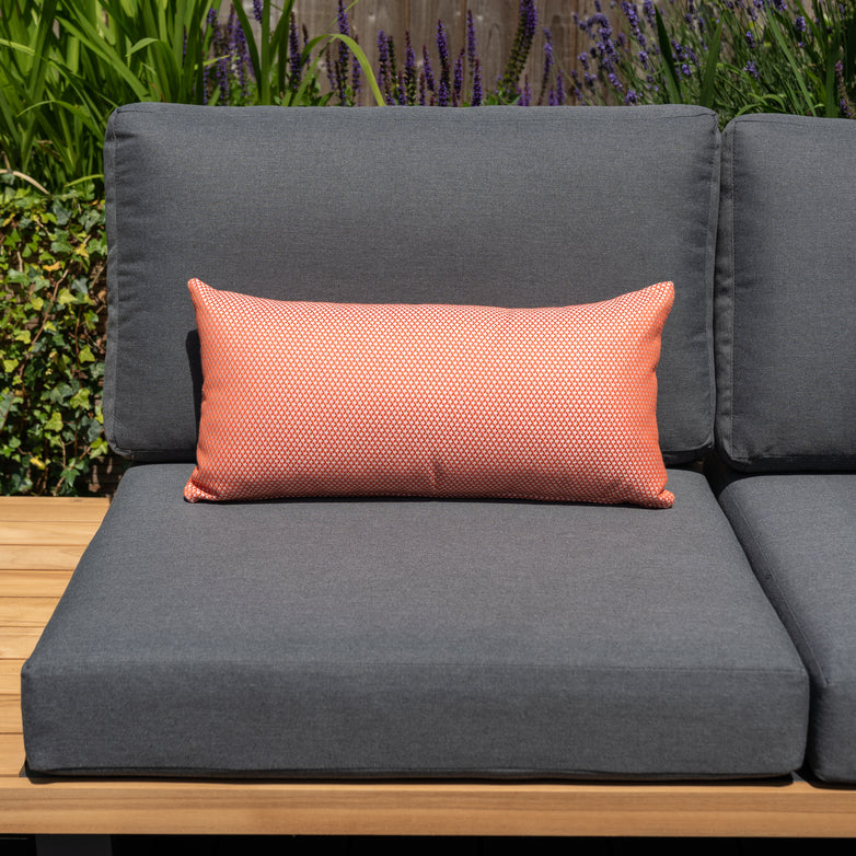 Gray outdoor sofa with a long, rectangular Agora Scala Coral Bolster Scatter Cushion - 60cm x 30cm made of solution-dyed acrylic against a backdrop of greenery and purple flowers.
