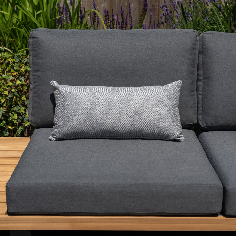 Outdoor gray cushioned sofa with an Agora Senda Ceniza Bolster Scatter Cushion (60cm x 30cm), placed on a wooden deck with green plants in the background.