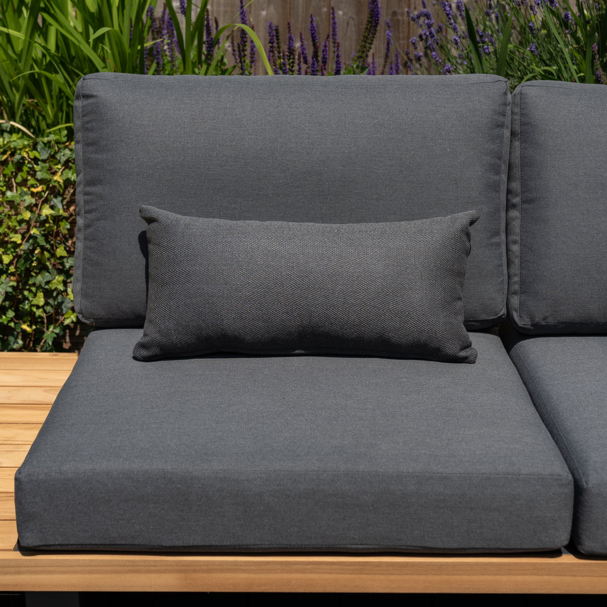 The Agora Esquire Mica Bolster Scatter Cushions (60cm x 30cm) are set on a wooden bench, with green plants and purple flowers adding to the backdrop. Crafted from solution-dyed acrylic, these outdoor scatter cushions bring both comfort and durability to your garden setup.