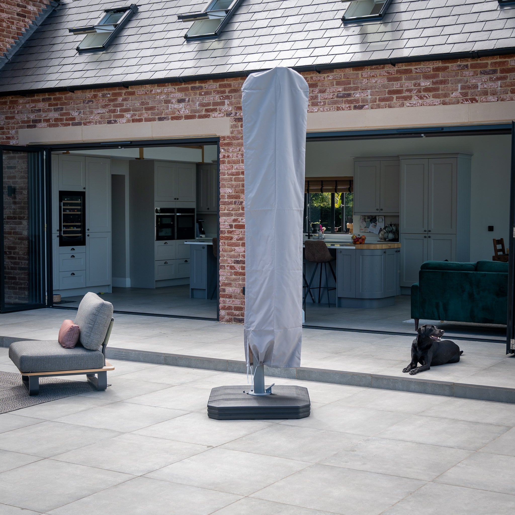 On a terrace in front of a brick house, there's a covered patio heater next to a grey chair and an elegant Pallas 4m x 3m Rectangular Cantilever Parasol with LED Lighting in Charcoal, accompanied by a covered barbecue and a black dog resting nearby.