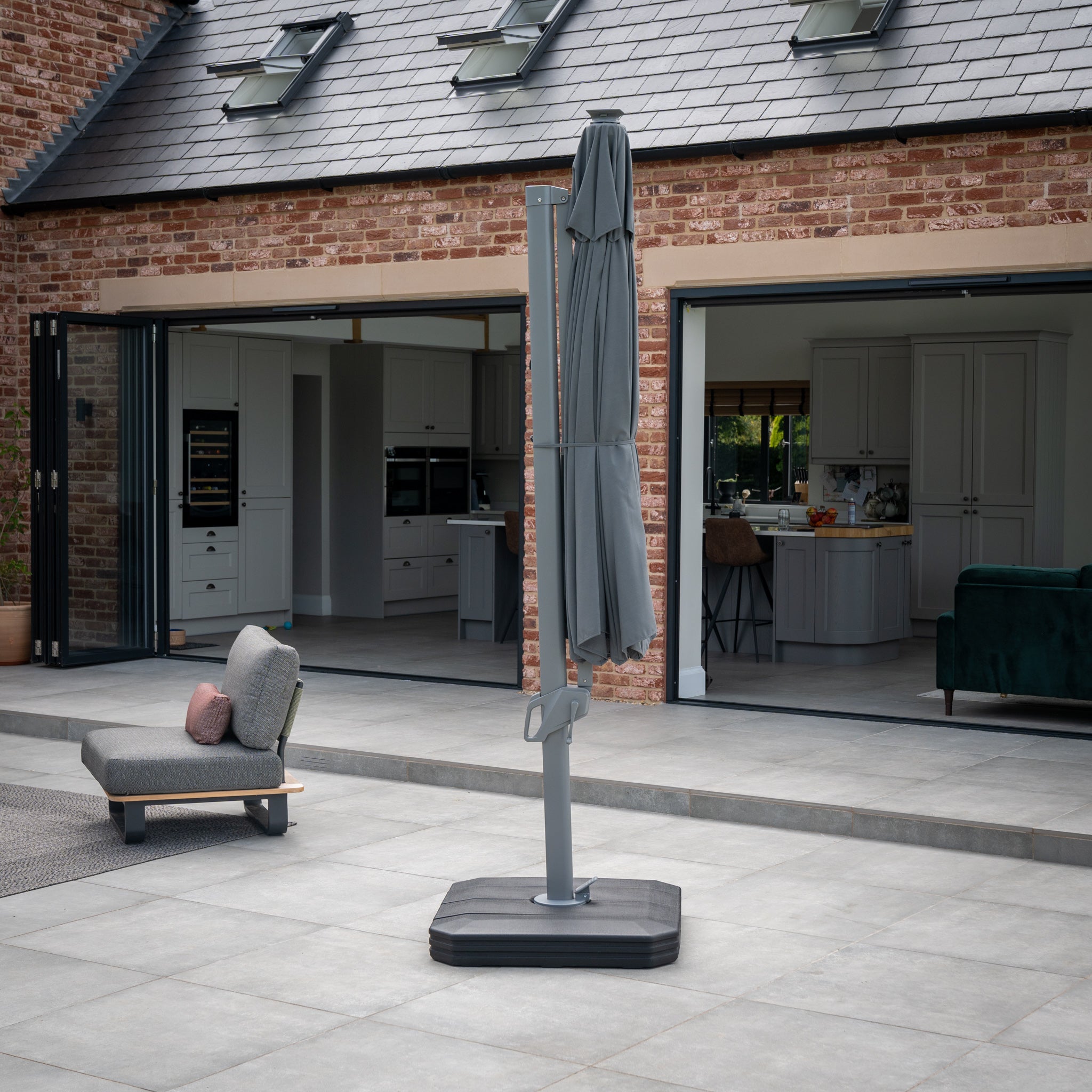 A closed Ares 3.5m Round Cantilever Parasol with Solar-powered LED Lights in Charcoal stands elegantly on a modern backyard patio, which features glass doors leading to a spacious kitchen and living area.
