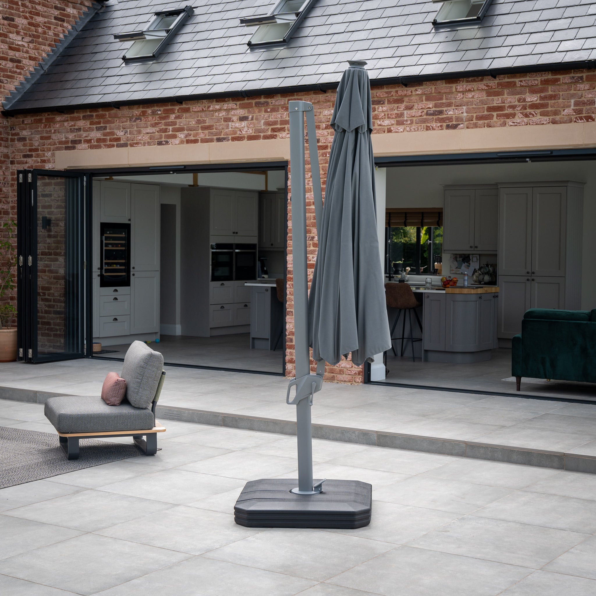 A charcoal Ares 3.5m round cantilever parasol with solar-powered LED lights is closed and set in a modern backyard patio near an open kitchen area with brick and tile elements.