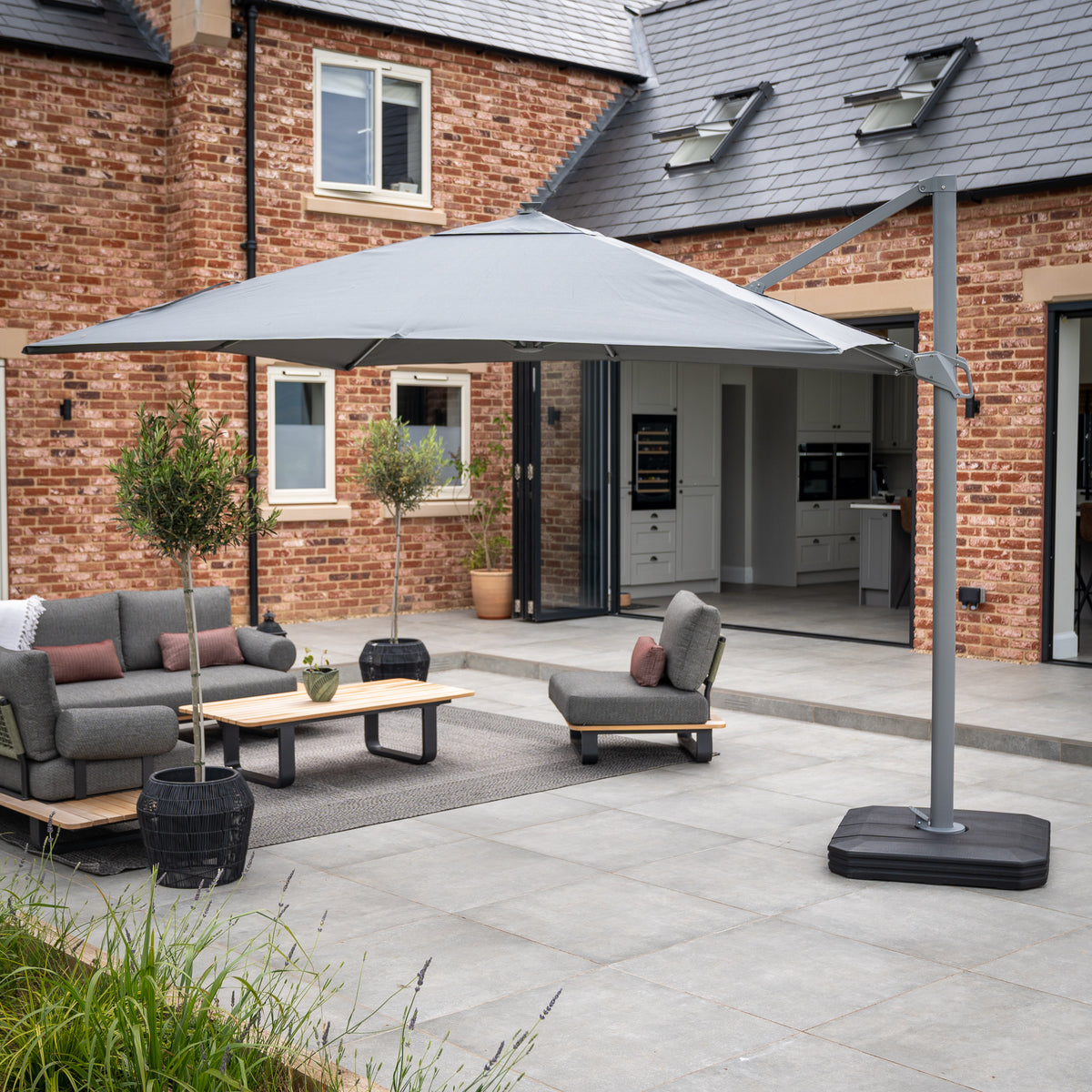 Modern patio with grey furnishings, a large Ares 3m Square Cantilever Parasol in Charcoal featuring an UV50+ canopy and solar-powered LED lights, and a brick house in the background.
