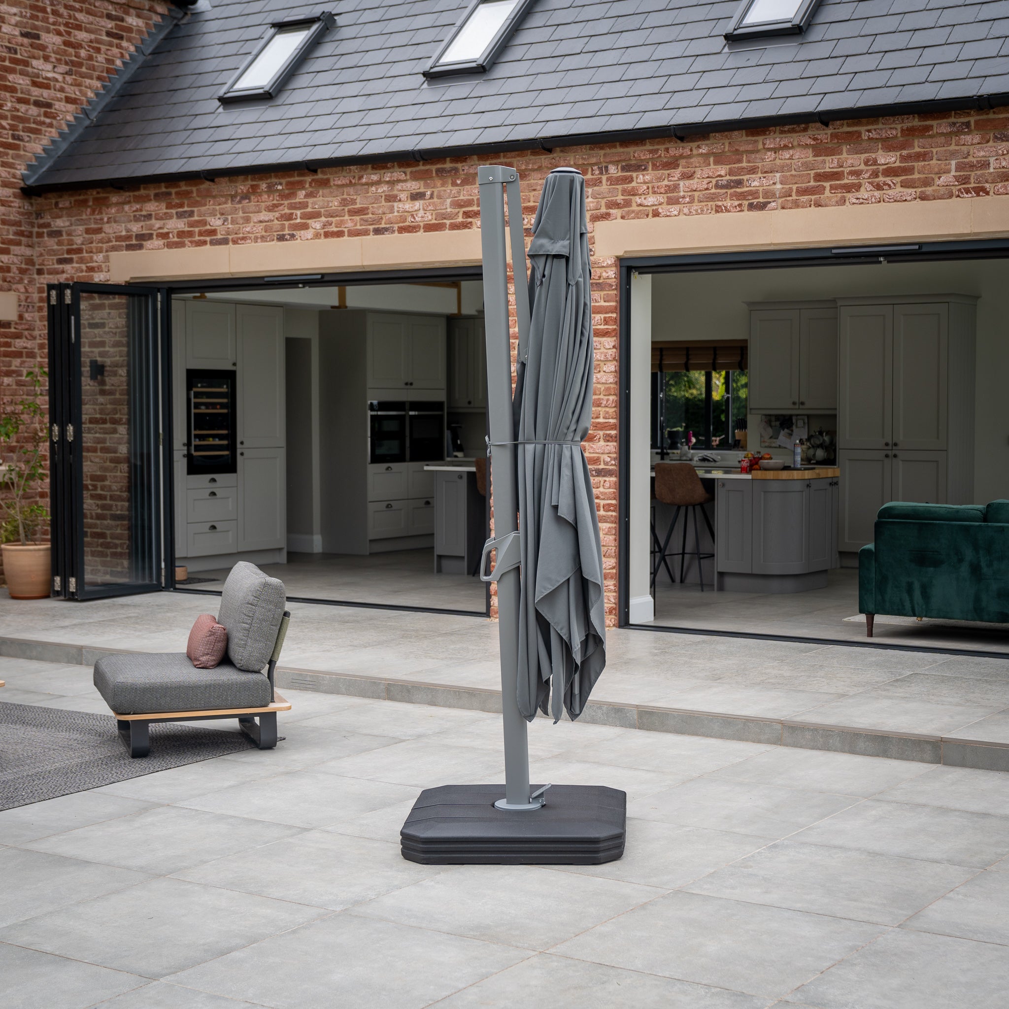 Patio with an Eros 3.3m x 2.4m Rectangular Cantilever Parasol in Charcoal featuring a closed UV50+ canopy, a lounge chair, and a modern brick house in the background with open sliding doors.