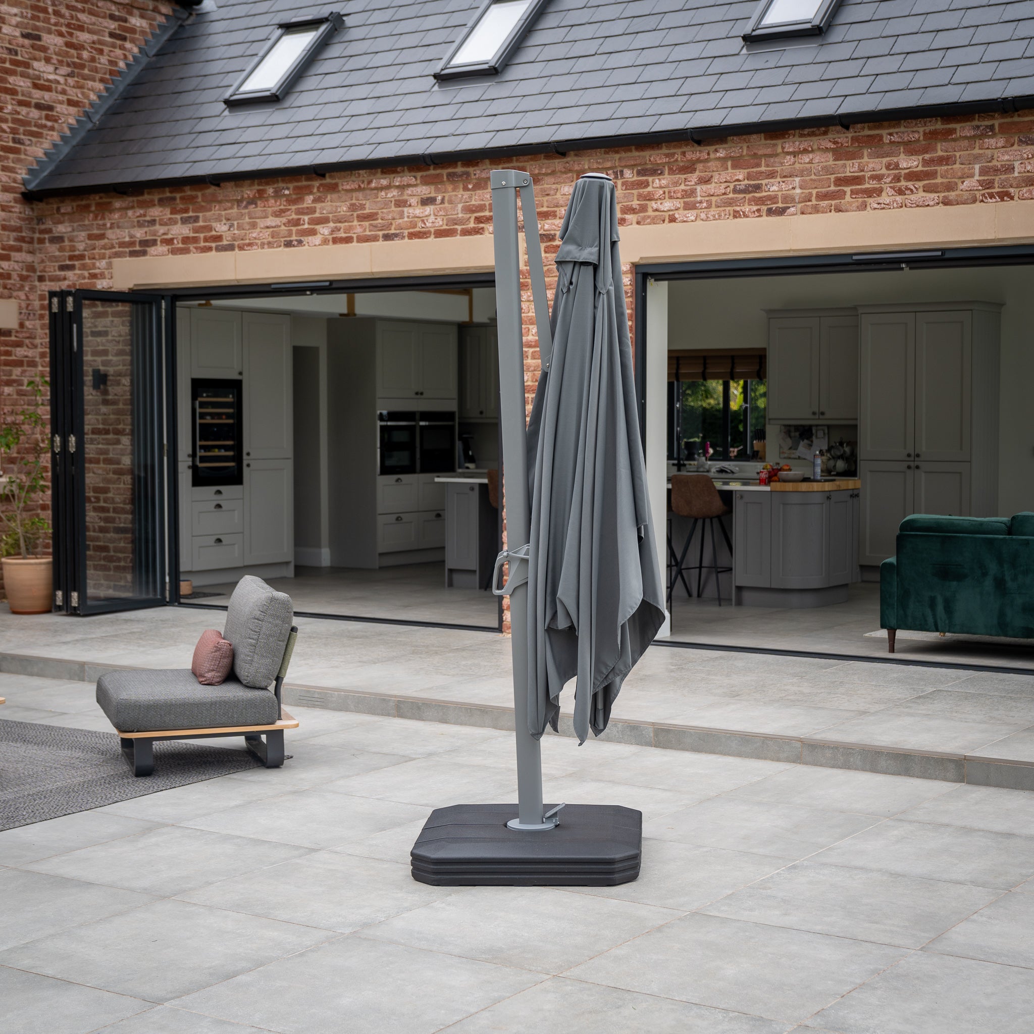 The Eros 3.3m x 2.4m Rectangular Cantilever Parasol in Charcoal stands closed on a tiled outdoor area in front of a modern house with an open sliding glass door, its UV50+ canopy blending seamlessly with the elegant surroundings.