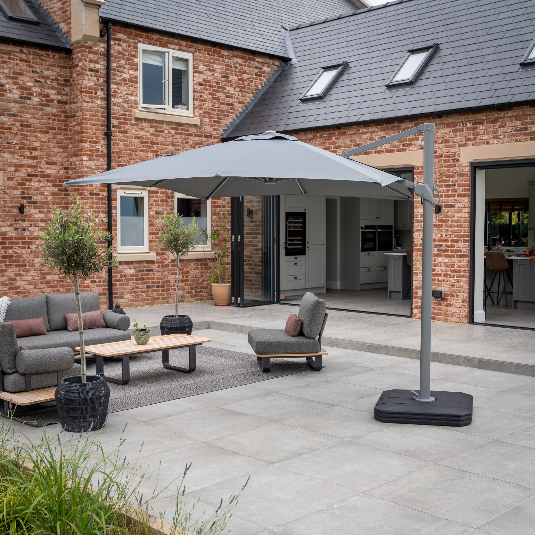 Modern patio with gray furniture and an Eros 3.3m x 2.4m Rectangular Cantilever Parasol in Charcoal, set against a brick house. Open doors lead to the kitchen.