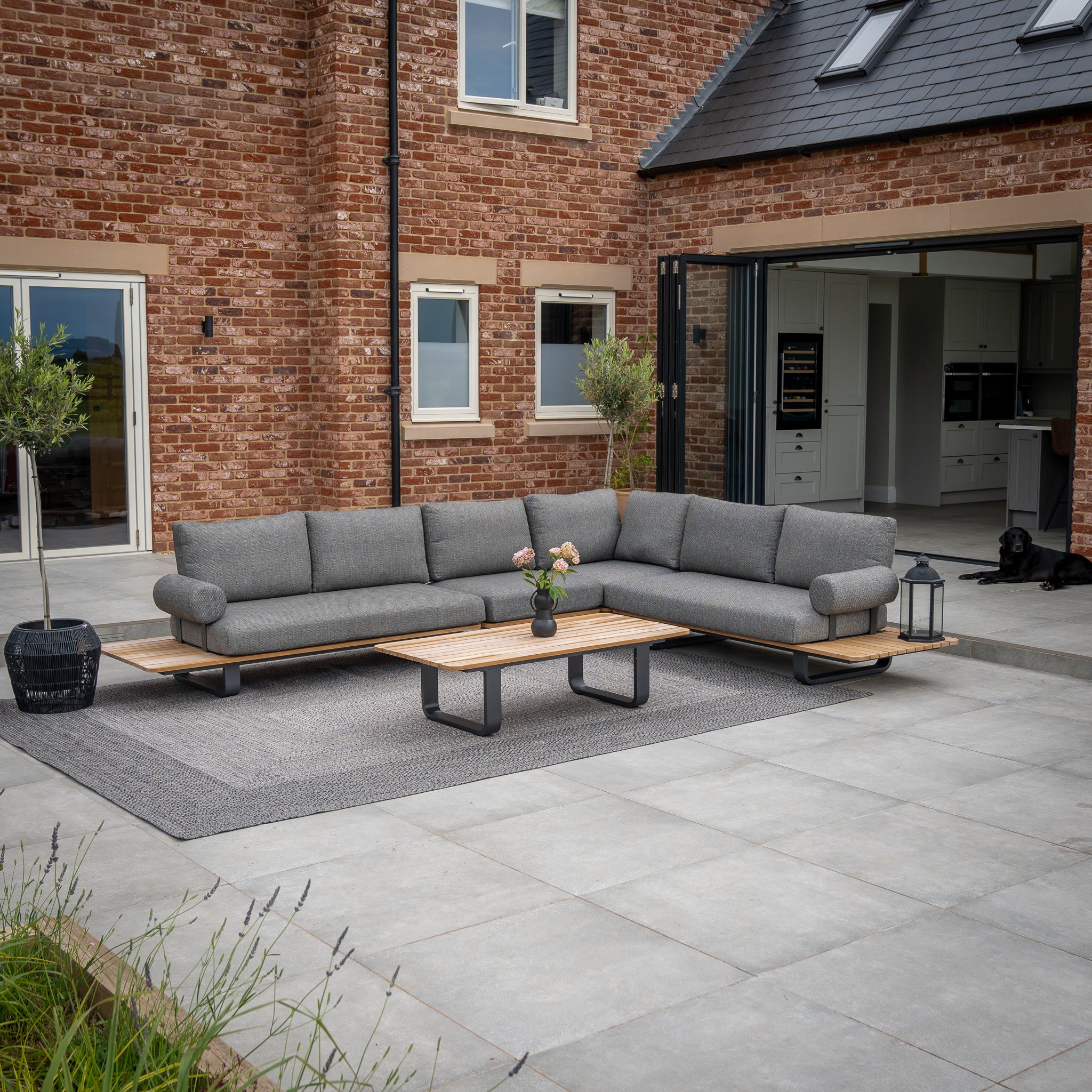 Modern outdoor patio featuring a cozy Bay Large Corner Group Set with 4 Waist Pillows in Olive Green and a Teak Table, complemented by potted plants on a large rug, right outside a charming brick house.