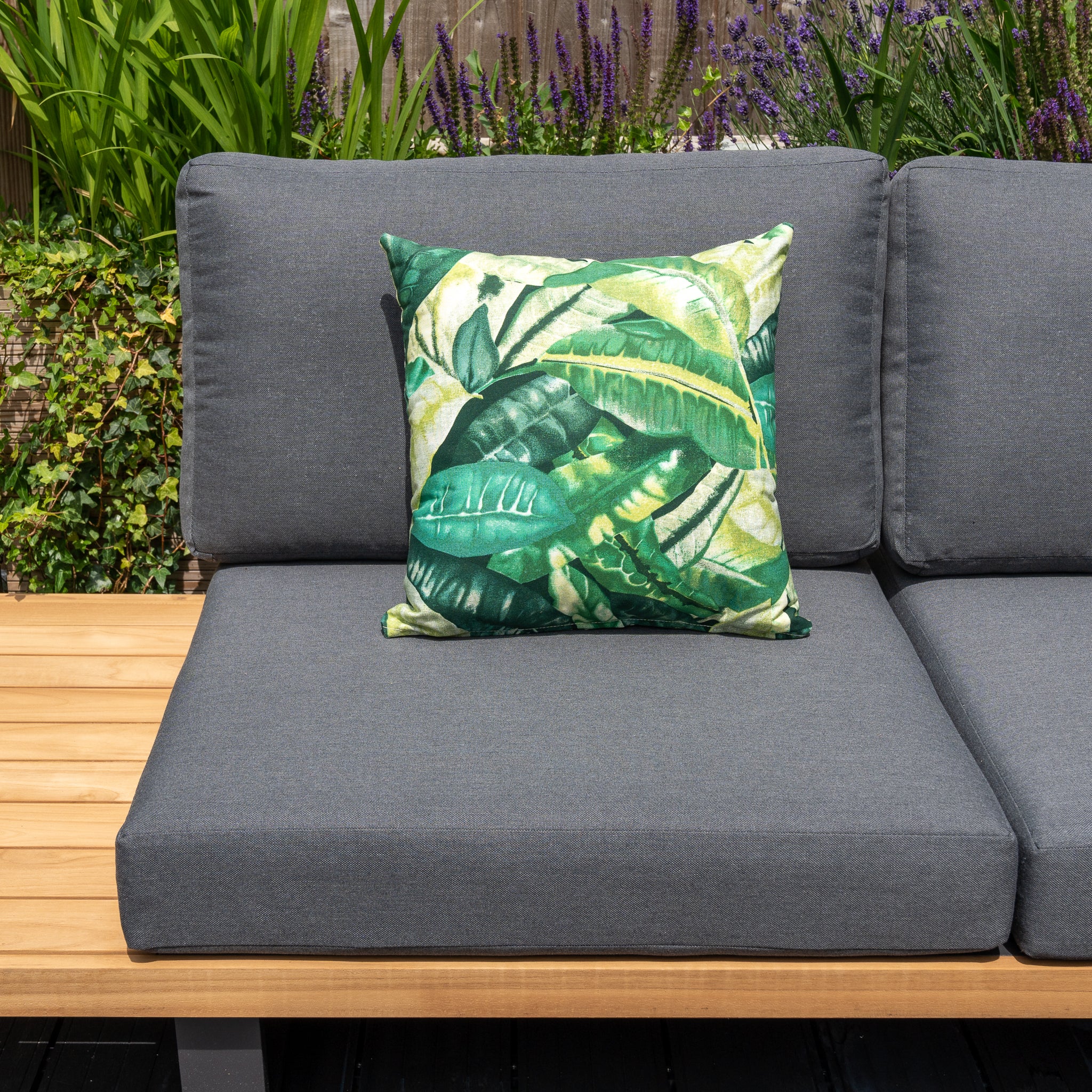 A gray outdoor sofa with a wooden side table, featuring the Acrisol Amazonia Verde Medium Scatter Cushion - 45cm x 45cm, adorned with a green leaf pattern and crafted from solution-dyed acrylic, set against a garden backdrop.
