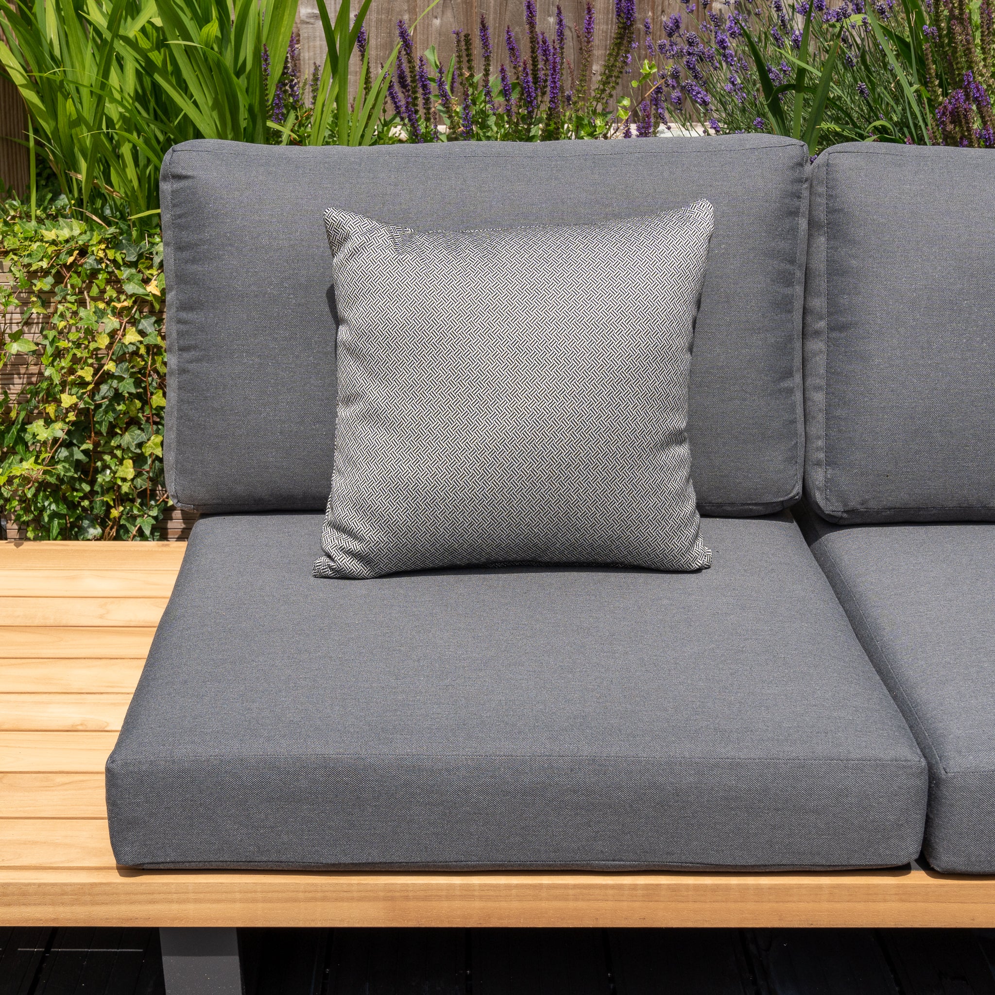 A grey outdoor sofa with a matching cushion, complemented by vibrant green plants and lavender flowers, is perfect for relaxing. Enhance it with the Agora Vimini Pirita Medium Scatter Cushion - 45cm x 45cm, made from durable solution-dyed acrylic to maintain color vibrancy.
