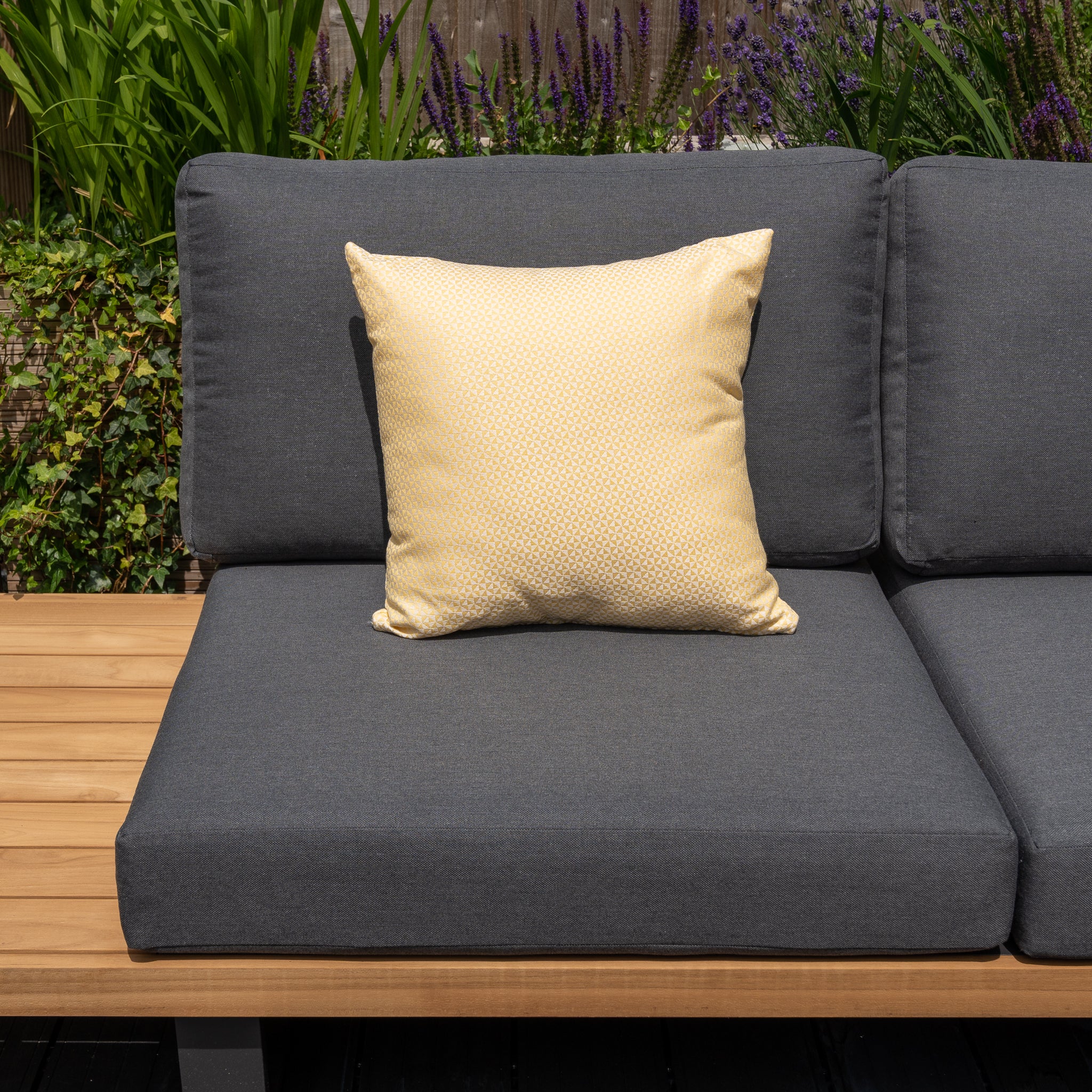 A gray outdoor couch with a yellow cushion on it, surrounded by greenery and purple flowers in the background, thoughtfully complemented by Acrisol Helix Trigo Medium Scatter Cushions - 45cm x 45cm.