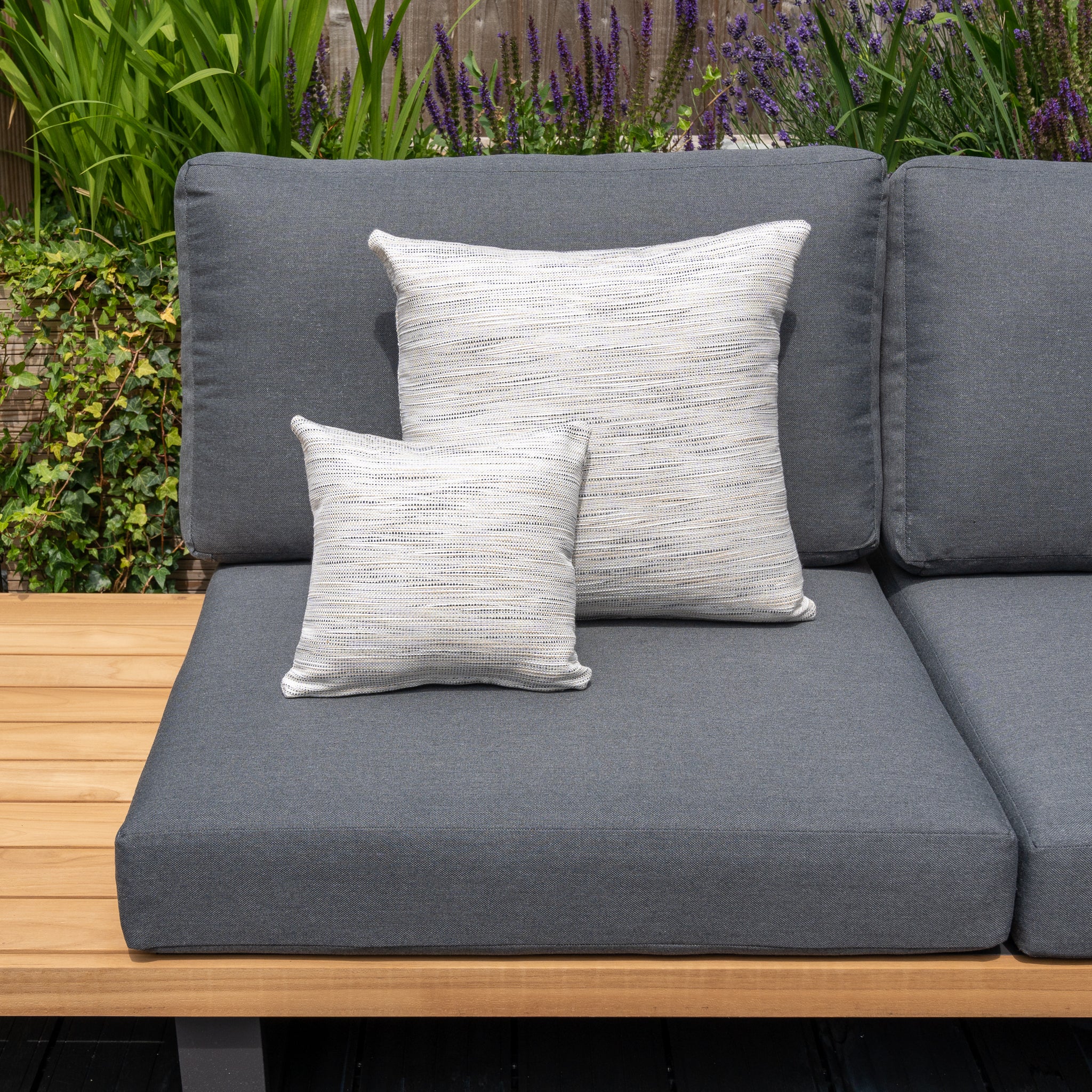 Outdoor patio sofa with grey, solution-dyed acrylic cushions, two Agora Texture Ugo Small Scatter Cushions (25cm x 25cm), and wooden armrests, surrounded by greenery and flowers.