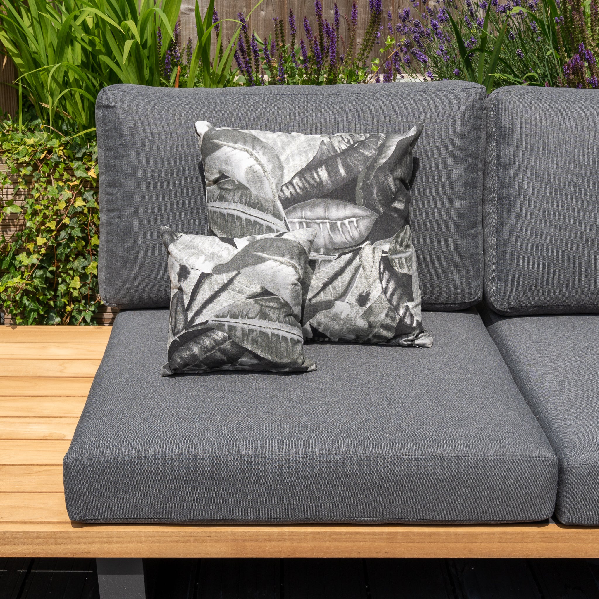 Outdoor grey sofa with two Acrisol Amazonia Gris small scatter cushions (25m x 25cm), set on a wooden deck surrounded by lush green and purple plants.
