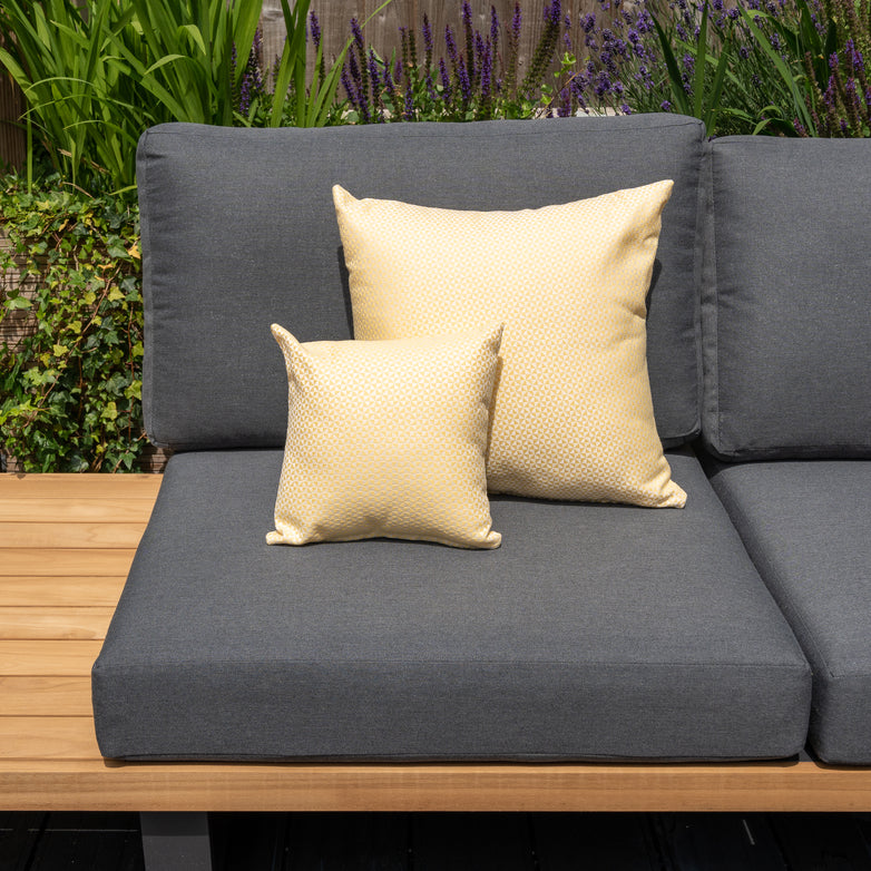 Grey outdoor couch adorned with two yellow outdoor scatter cushions, including the Acrisol Helix Trigo Small Scatter Cushion (25cm x 25cm), set against a backdrop of lush green foliage.