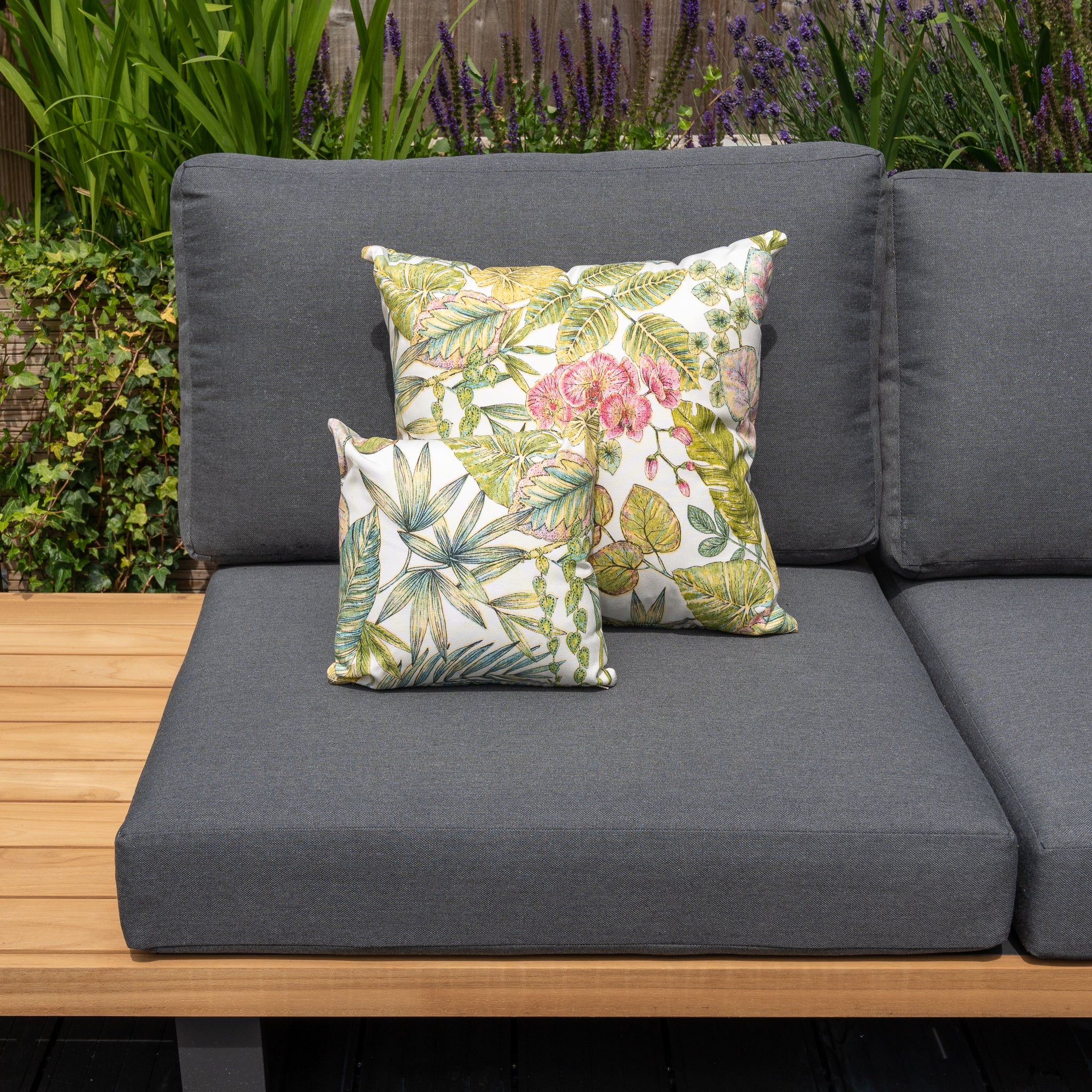 Gray outdoor sofa with floral patterned throw pillows and a wooden side table, surrounded by green plants and flowers. The addition of Acrisol Bouquet Multi Small Scatter Cushion - 25m x 25cm in solution-dyed acrylic ensures long-lasting, vibrant colors.