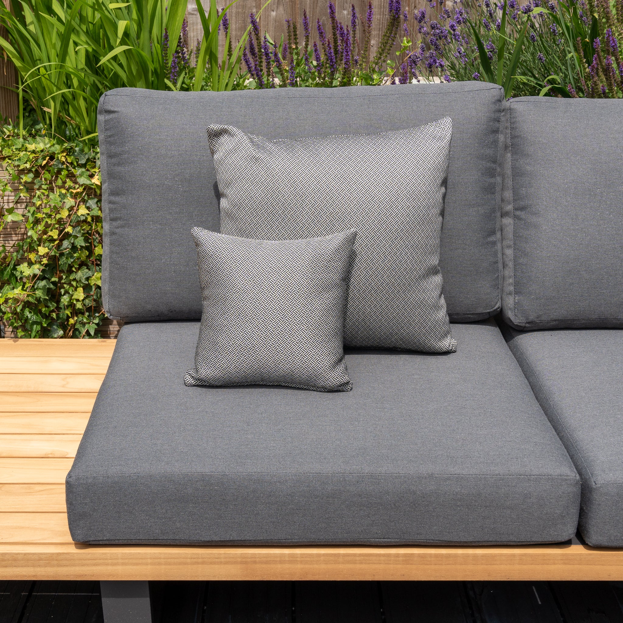 A grey outdoor sofa, adorned with Harbour Lifestyle cushions and a mix of large and small pillows including the Agora Vimini Pirita Small Scatter Cushion (25cm x 25cm), sits against a garden backdrop with vibrant green plants and purple flowers. The solution-dyed acrylic fabric ensures lasting color and durability.
