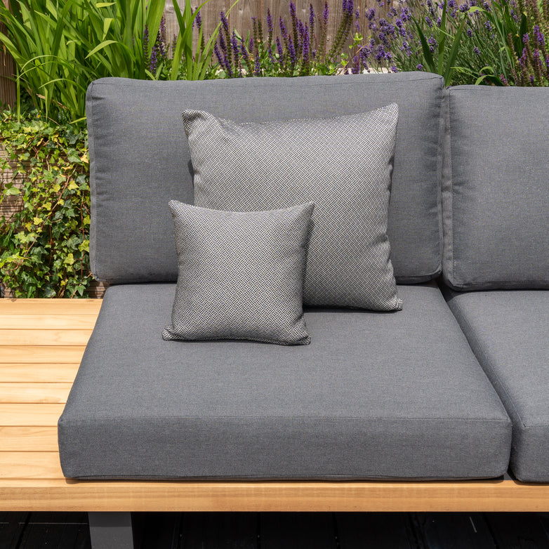 A grey outdoor sofa, adorned with Harbour Lifestyle cushions and a mix of large and small pillows including the Agora Vimini Pirita Small Scatter Cushion (25cm x 25cm), sits against a garden backdrop with vibrant green plants and purple flowers. The solution-dyed acrylic fabric ensures lasting color and durability.