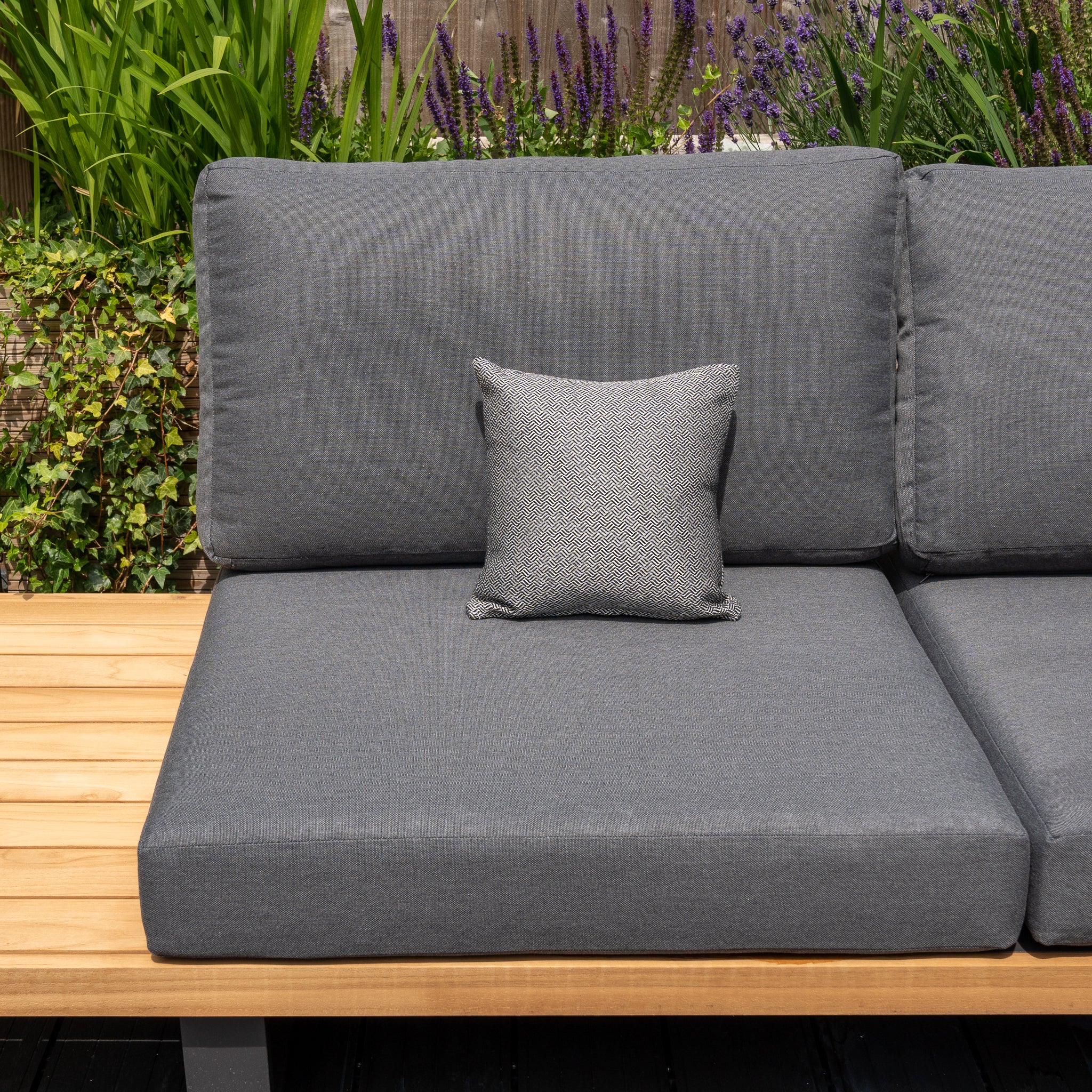 A gray outdoor sofa with a small gray Agora Vimini Pirita Small Scatter Cushion (25cm x 25cm), featuring Harbour Lifestyle cushions, set against a wooden base with green and purple plants in the background.