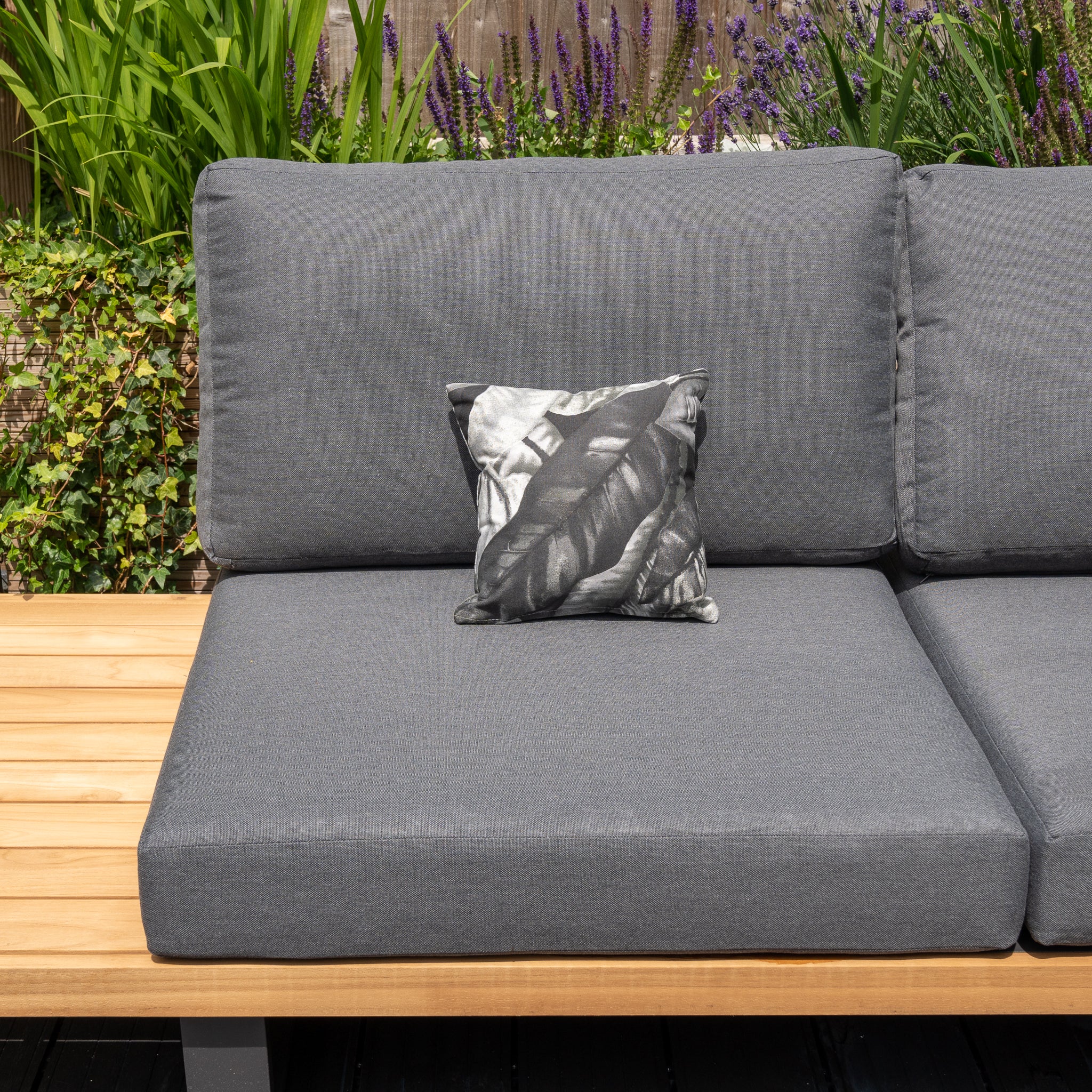 Outdoor gray cushioned bench with a black and white leaf-pattern pillow, surrounded by green plants and purple flowers. Enhance your setup with the Acrisol Amazonia Gris Small Scatter Cushion – 25cm x 25cm, made from solution-dyed acrylic for superior durability.