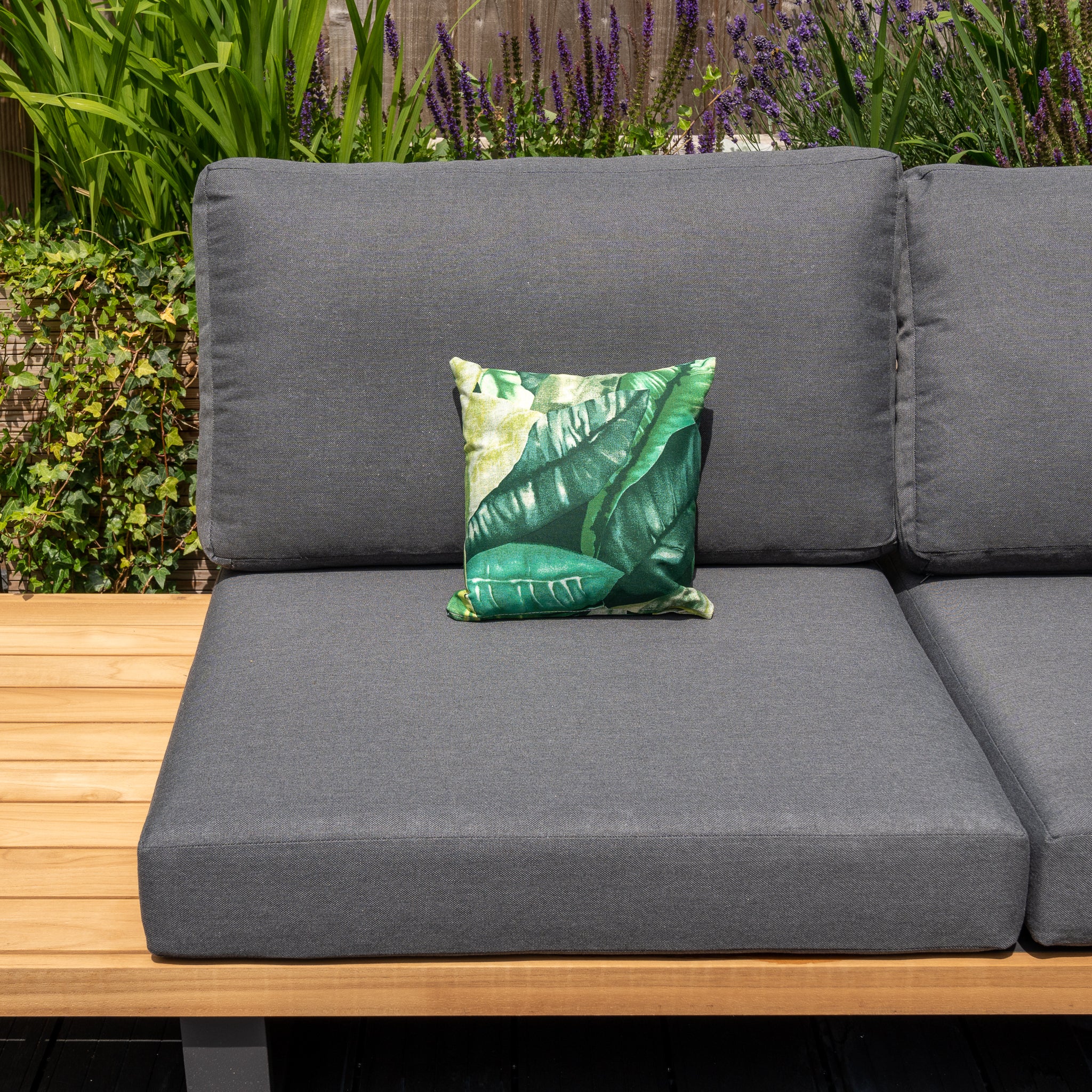 A gray outdoor sofa with an Acrisol Amazonia Verde Small Scatter Cushion - 25cm x 25cm, showcasing a sophisticated green leaf pattern, is placed elegantly against a backdrop of lush plants and flowers. The solution-dyed acrylic material ensures lasting color and durability.