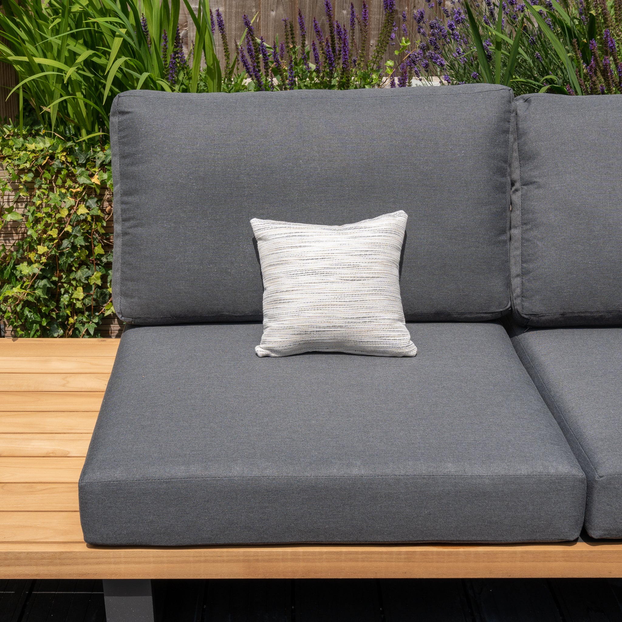 A gray outdoor sofa with an Agora Texture Ugo Small Scatter Cushion - 25cm x 25cm, placed on a wooden deck with green plants and purple flowers in the background, benefits from the durability of solution-dyed acrylic fabric.