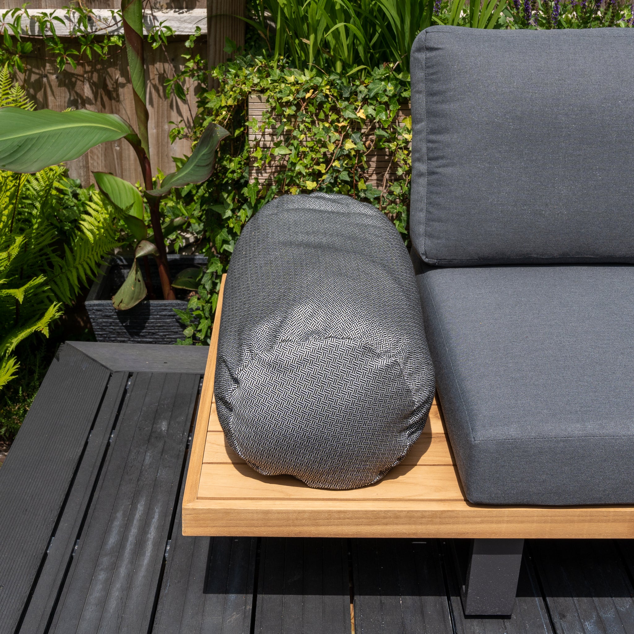 A gray outdoor sofa with a matching pillow on a wooden platform is surrounded by lush green plants and foliage. Complement it with the vibrant Agora Vimini Pirita Large Round Bolster Floor Cushion - 72cm x 35cm and keep everything pristine by using a convenient cushion storage bag when not in use.