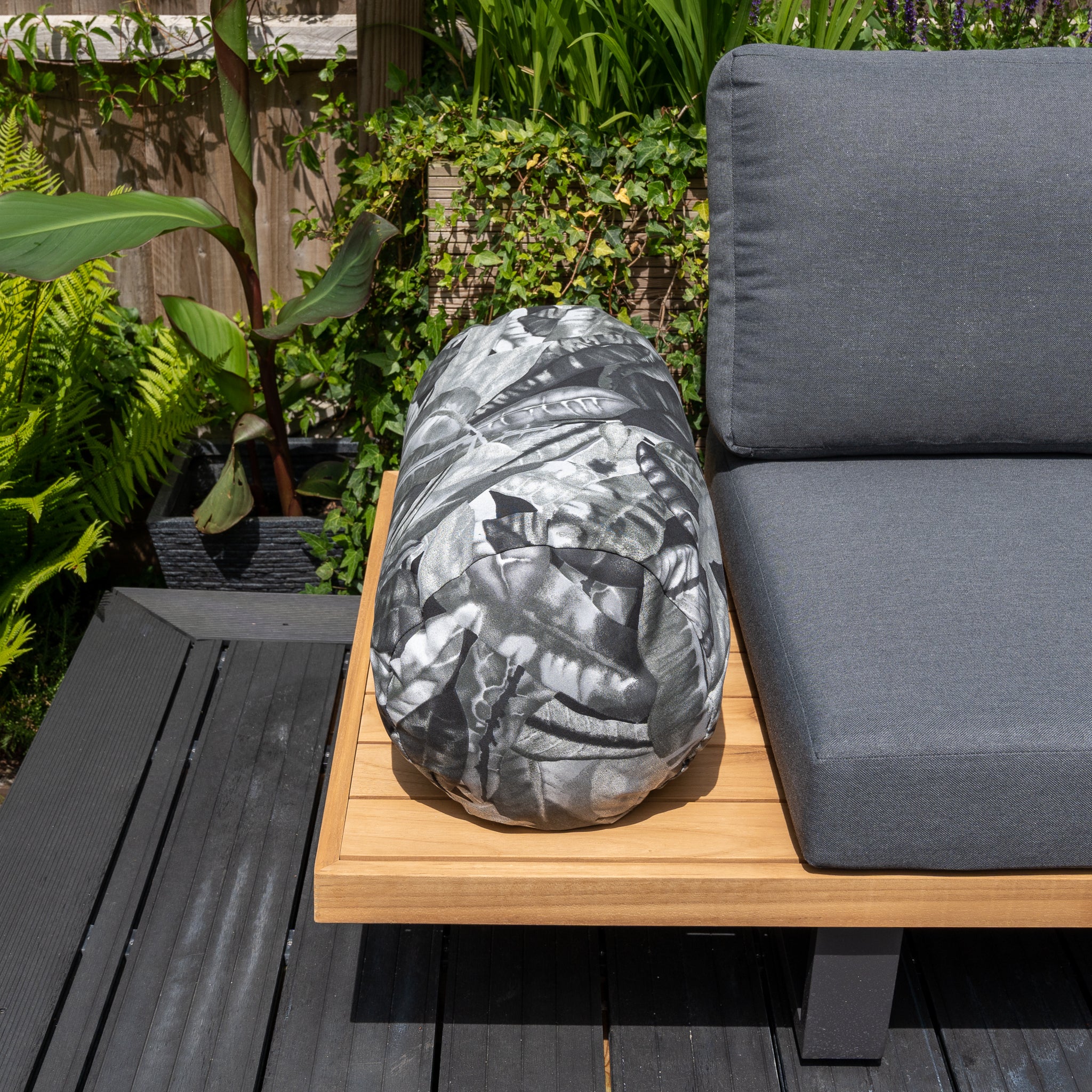 Outdoor seating featuring the Acrisol Amazonia Gris Large Round Bolster Floor Cushion in a lush garden setting. The gray cushion and cylindrical pillow boast a striking black and white leaf pattern, complemented by vibrant, durable solution-dyed acrylic fabrics ensuring lasting color amidst the greenery.