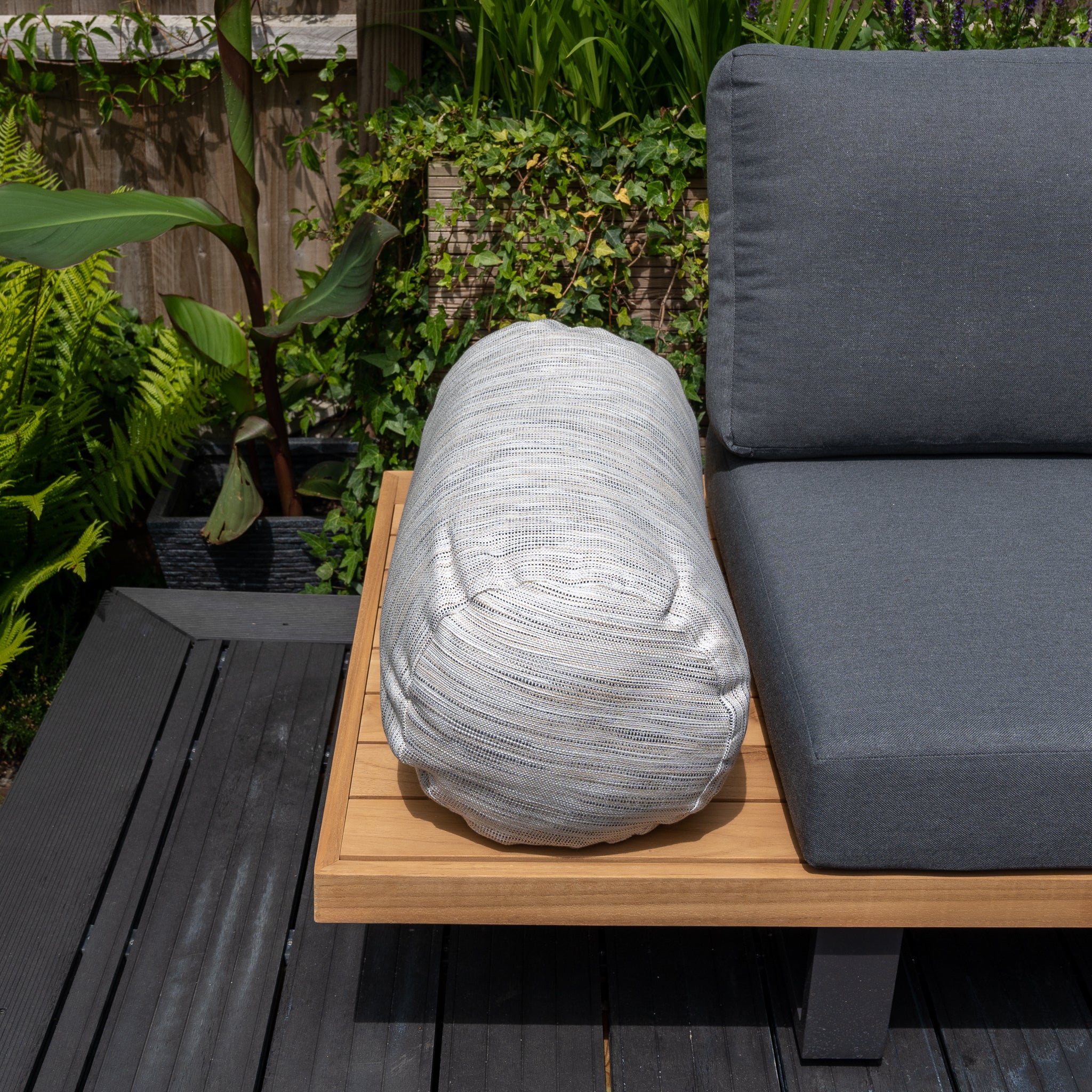 A modern outdoor lounge chair featuring gray solution-dyed acrylic cushions and the Agora Texture Ugo Large Round Bolster Floor Cushion (72cm x 35cm) on a wooden patio, surrounded by lush greenery for garden personalization.