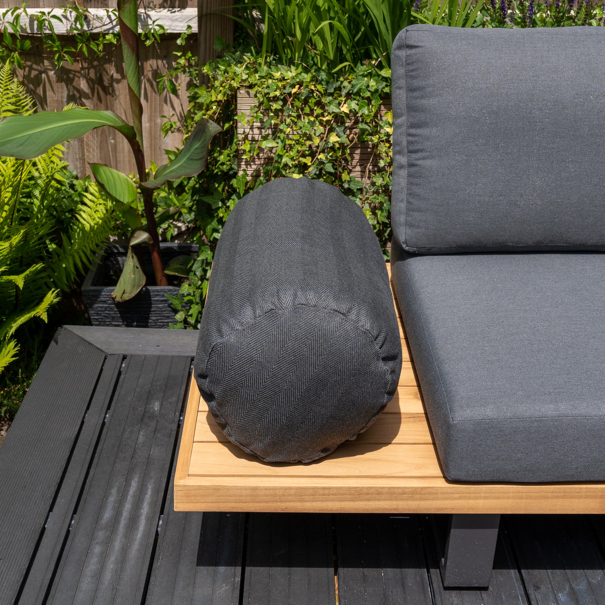 The Agora Esquire Mica Large Round Bolster Floor Cushion, measuring 72cm x 35cm, rests on a wooden bench with gray cushions, surrounded by plants in a garden setting and complemented by outdoor scatter cushions.