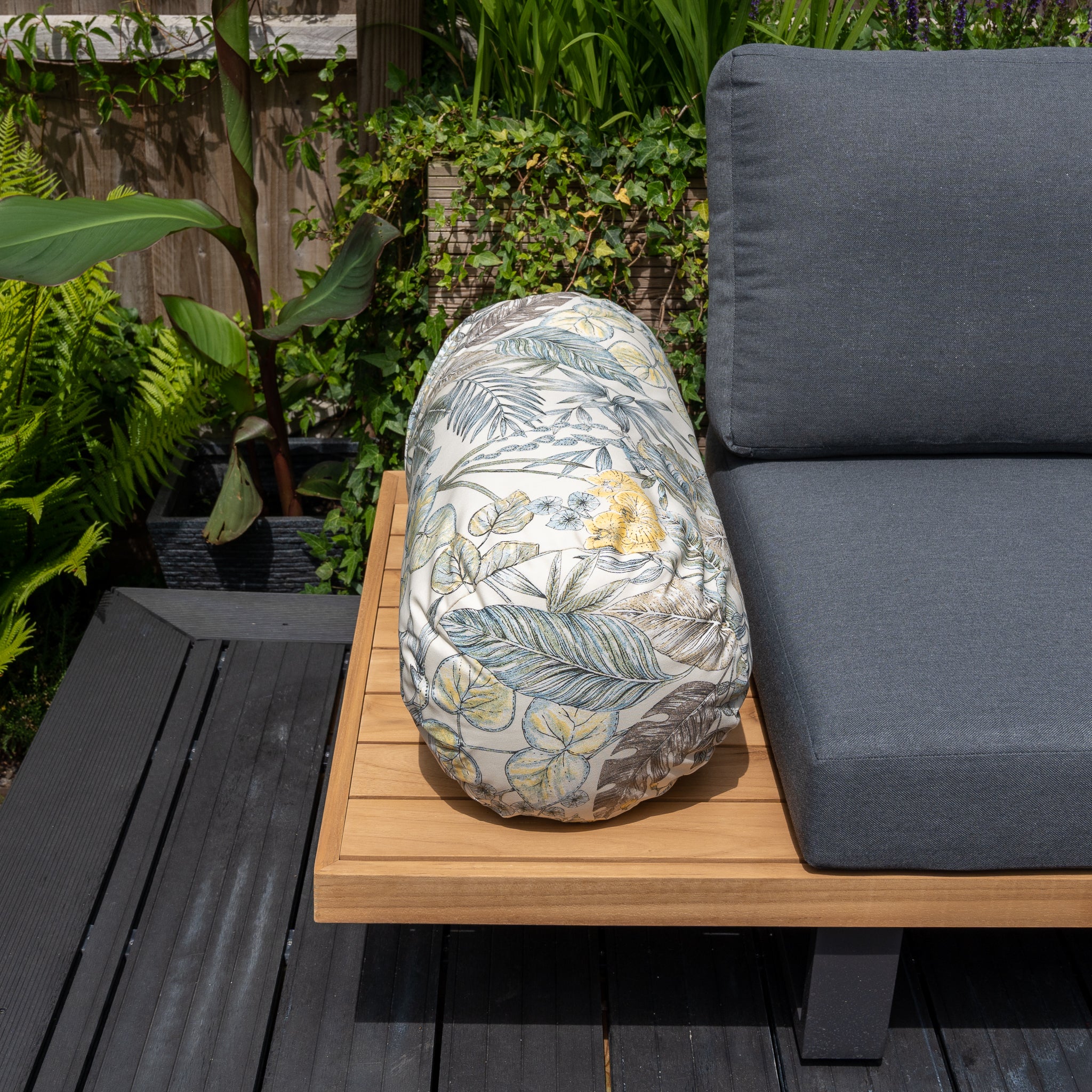 An Acrisol Bouquet Neutrals Large Round Bolster Floor Cushion (72cm x 35cm) with botanical prints sits on a wooden outdoor table next to a gray solution-dyed acrylic cushion.