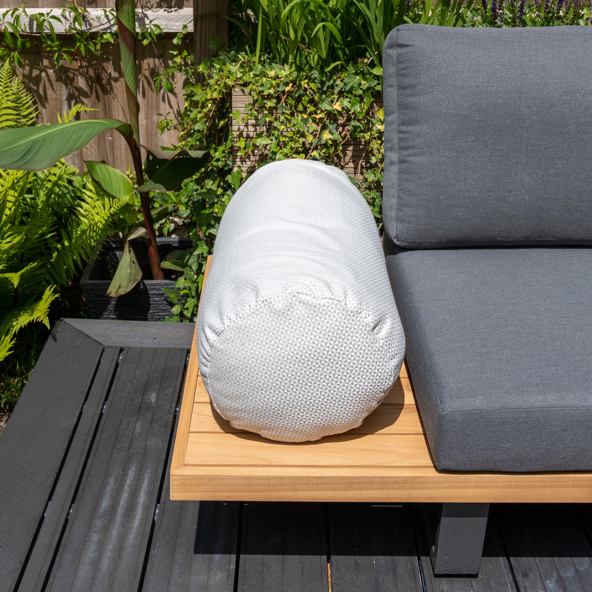 Acrisolid Helix Gris Claro Large Round Bolster Floor Cushion on a wooden outdoor bench next to gray outdoor scatter cushions, surrounded by lush green plants.