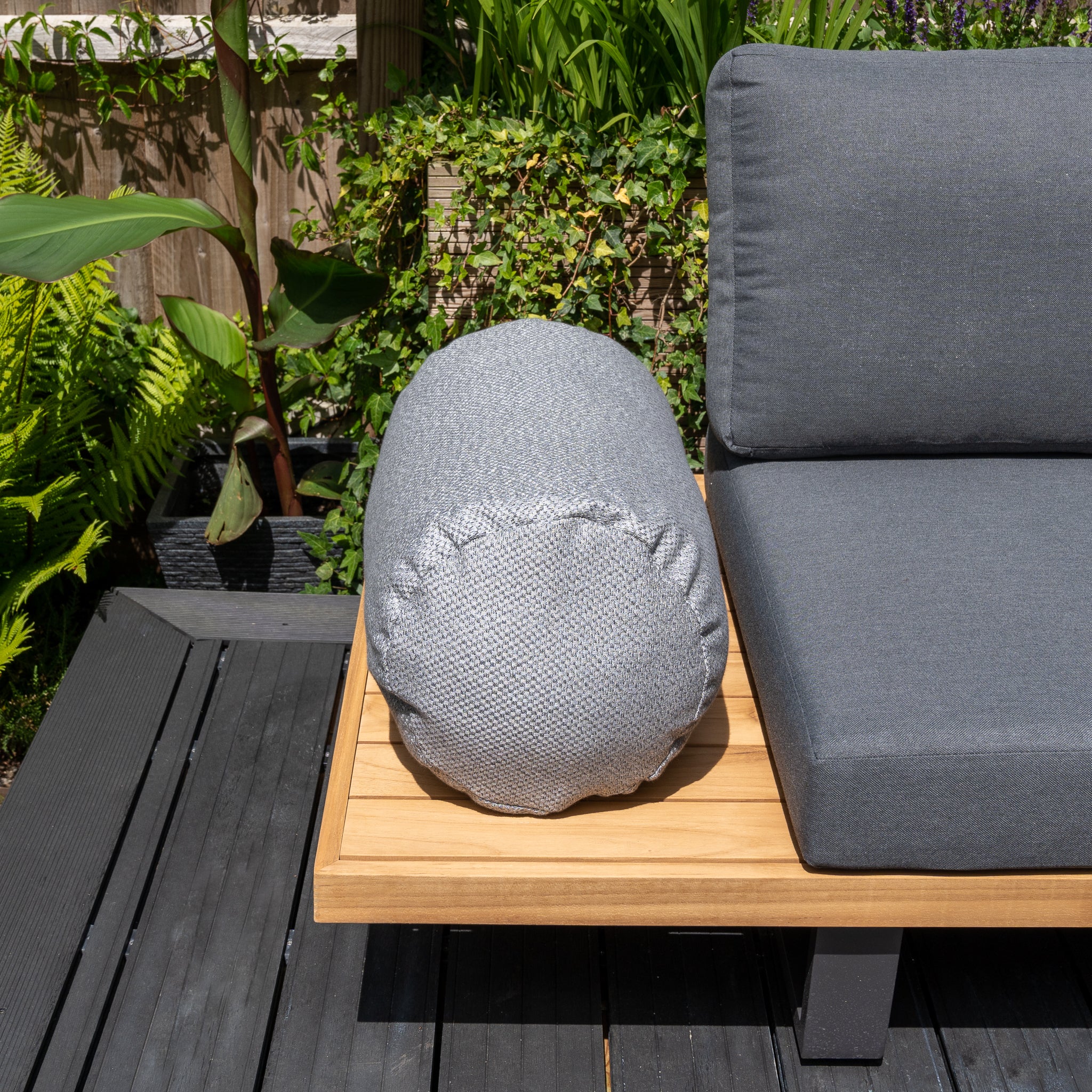 Agora Senda Ceniza Large Round Bolster Floor Cushion on wooden outdoor bench next to gray padded seating, adorned with outdoor scatter cushions and surrounded by green plants and a wooden fence. Keep everything pristine with the Golden Care Fabrics & Upholstery Care Kit.