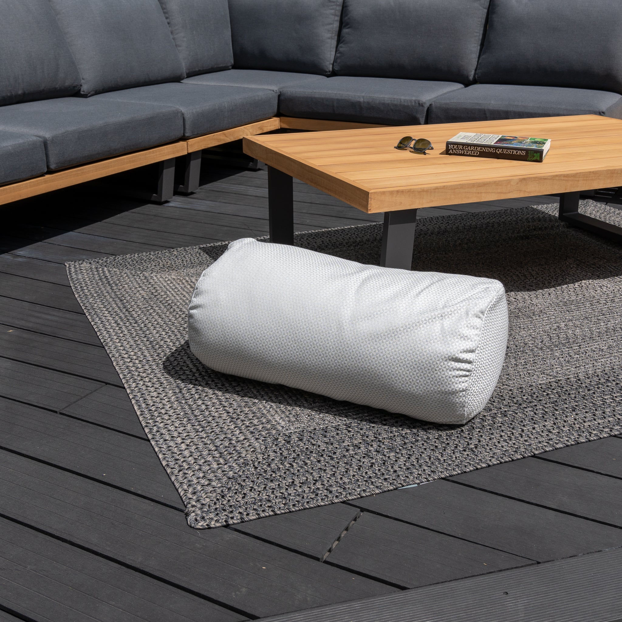 An Acrisol Helix Gris Claro Large Round Bolster Floor Cushion sits on a textured gray rug next to a wooden coffee table and a sectional outdoor sofa adorned with colorful outdoor scatter cushions.