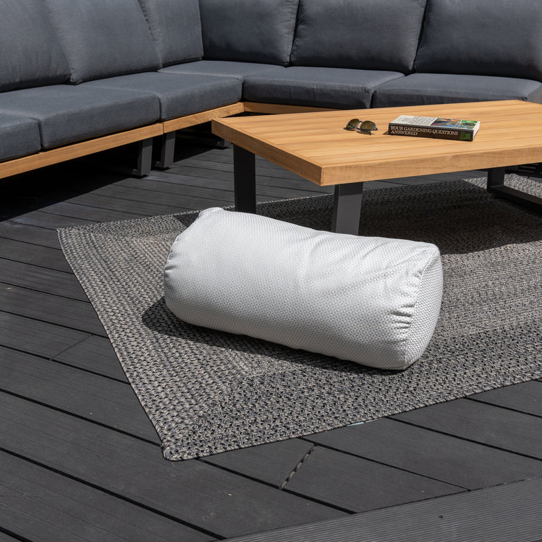 An Acrisol Helix Gris Claro Large Round Bolster Floor Cushion sits on a textured gray rug next to a wooden coffee table and a sectional outdoor sofa adorned with colorful outdoor scatter cushions.